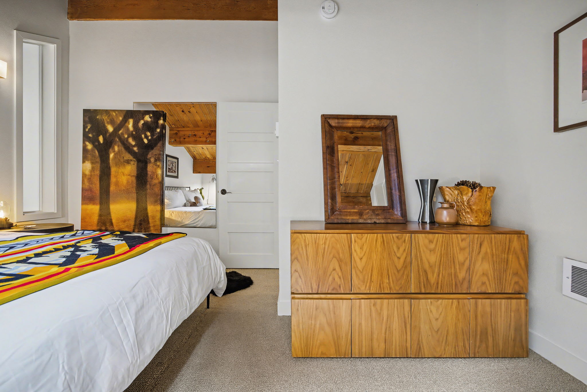 This bedroom view features a mid-century modern wooden dresser with multiple drawers, topped with decorative vases and a framed mirror. A large abstract painting leans against the wall, and a white door leads to another room, all under exposed wooden beams.