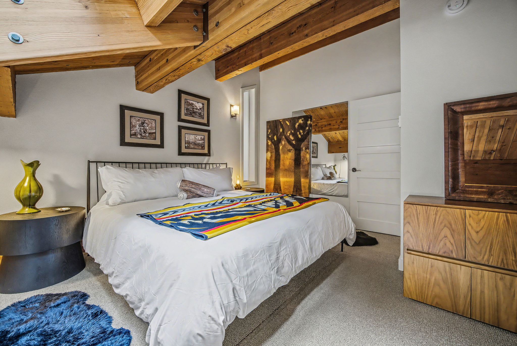 A cozy bedroom features a queen-sized bed with crisp white bedding and a vibrant, colorful patterned blanket, flanked by dark round nightstands. Exposed wooden beams accent the vaulted ceiling, and framed artwork adorns the white walls above a soft carpet.