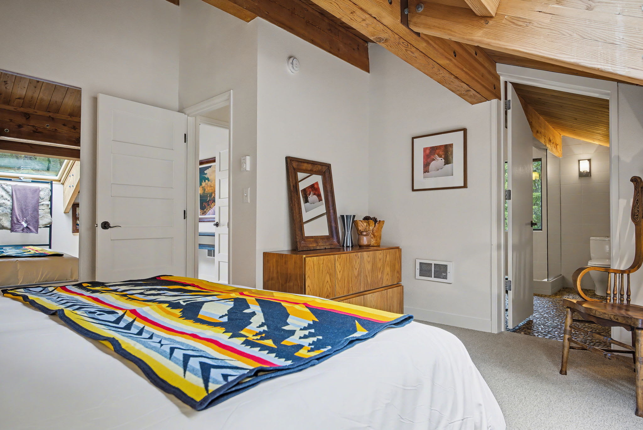 A bright bedroom with a white-linened bed and colorful blanket transitions into a bathroom through an open doorway, revealing dark pebble flooring. A wooden dresser with a mirror and a decorative chair are visible, all beneath a vaulted ceiling with exposed wooden beams.