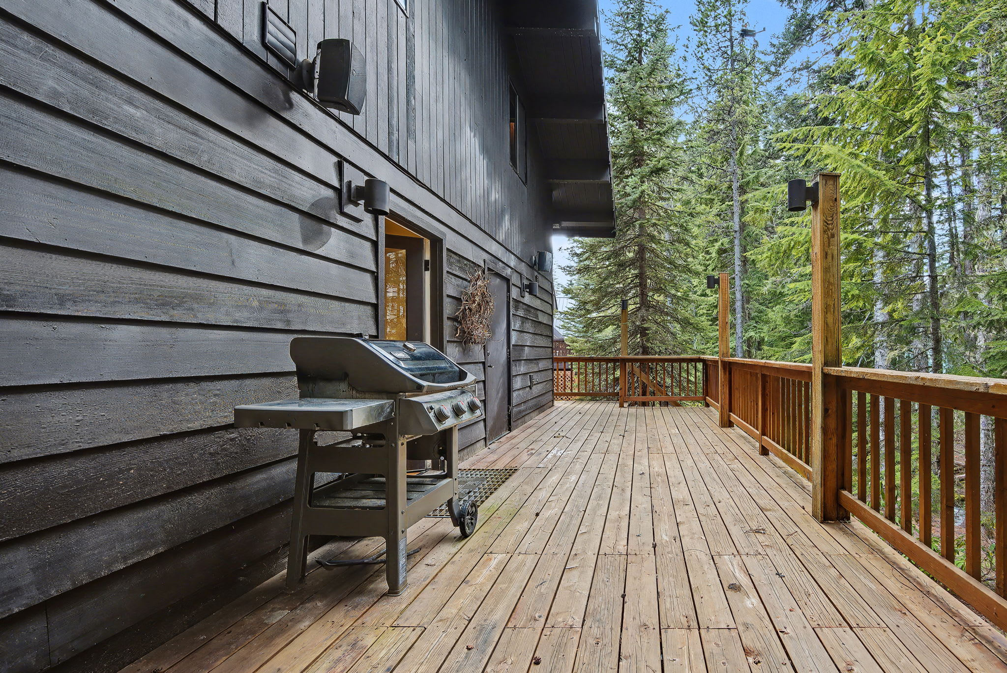 Expansive wooden deck with a gas grill, extending along the dark-stained exterior of the house, offering views of the surrounding tall evergreen trees.