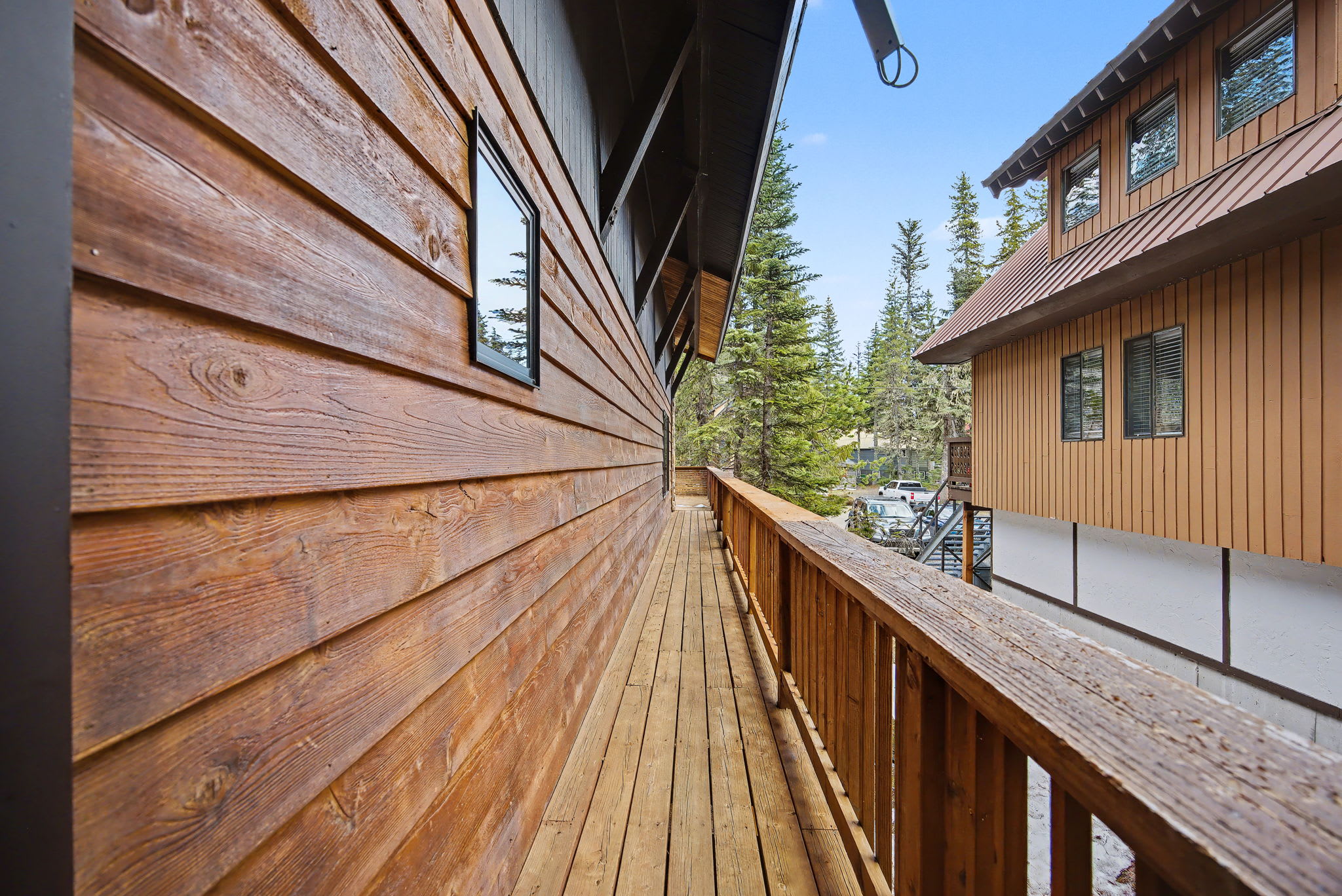 A narrow wooden balcony with a sturdy railing runs alongside a rustic wooden-sided building, offering a glimpse of another cabin and tall evergreen trees in the distance under a clear sky.