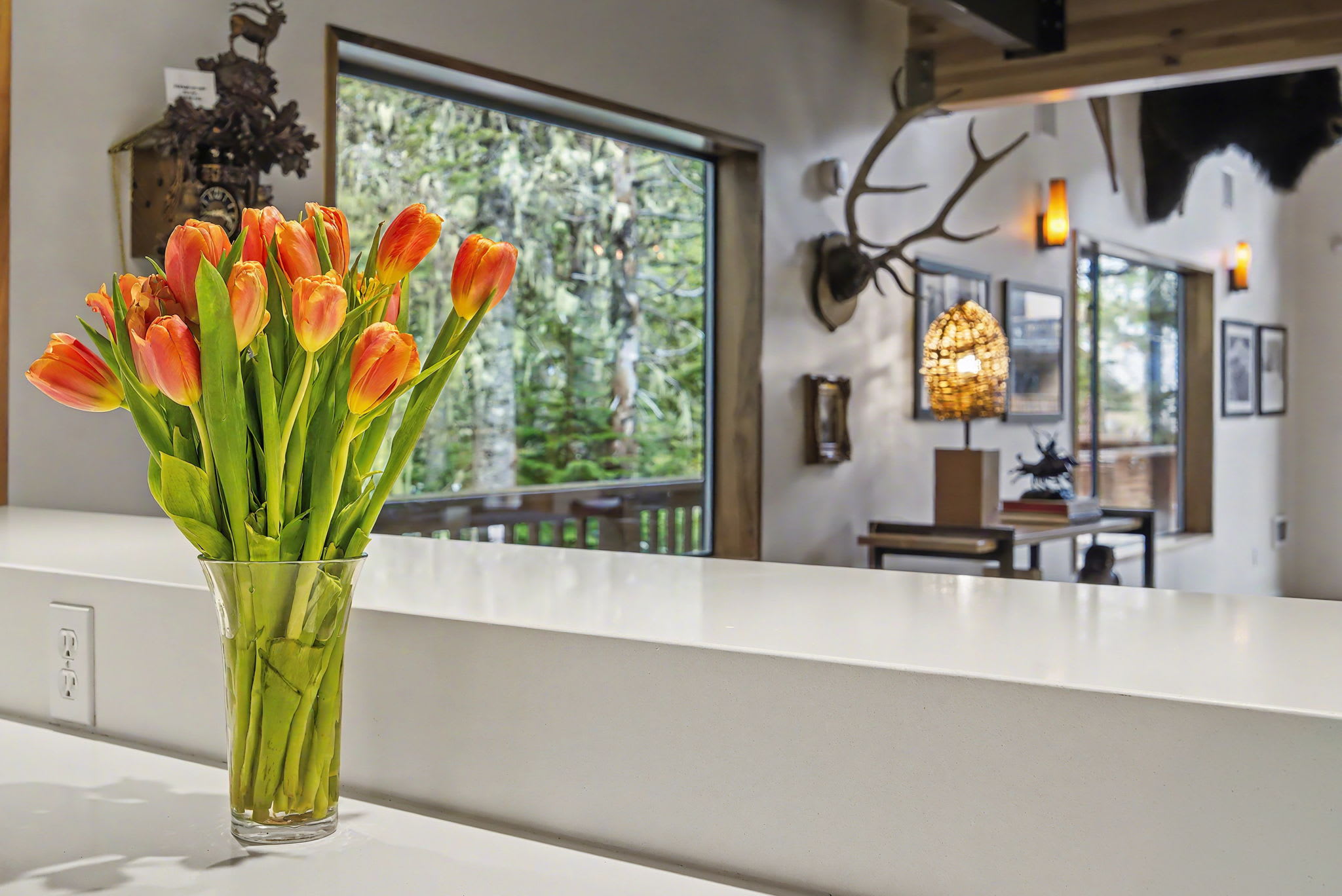 View from the kitchen bar area into the living space, featuring a vase of vibrant orange tulips on a white countertop. Large windows in the background offer views of lush greenery.