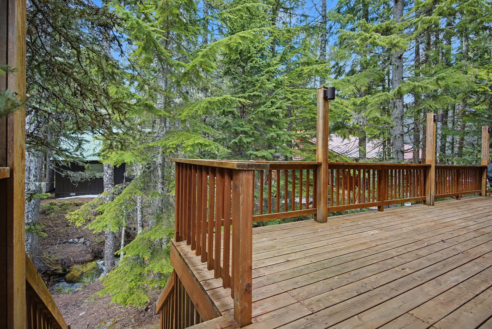 A spacious wooden deck with a sturdy wooden railing is nestled among dense evergreen trees, with stairs leading down and a glimpse of a stream or small waterfall below.