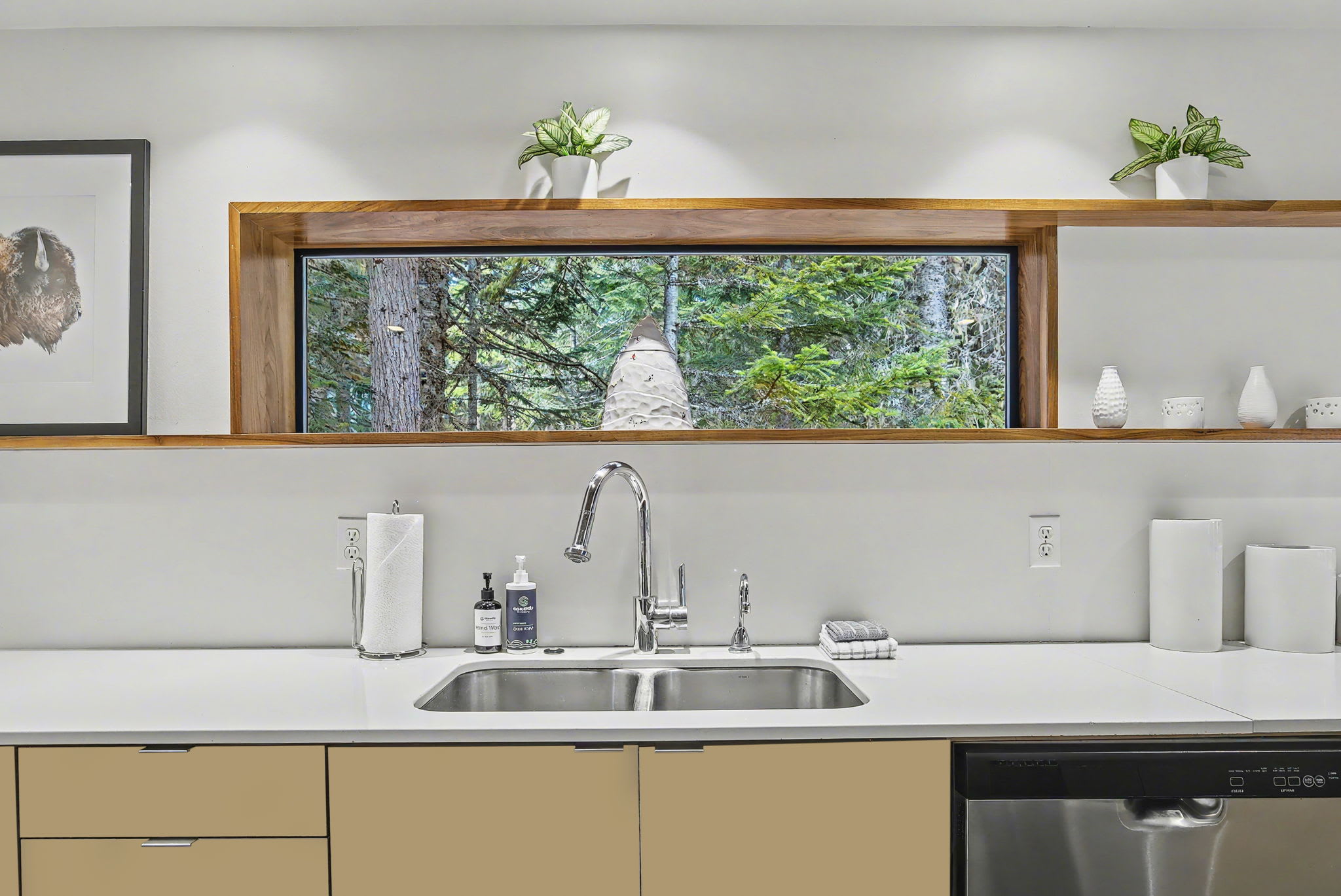 Kitchen sink area featuring a double stainless steel sink, chrome faucet, and white countertops. A horizontal window above the sink provides a serene view of the surrounding forest.