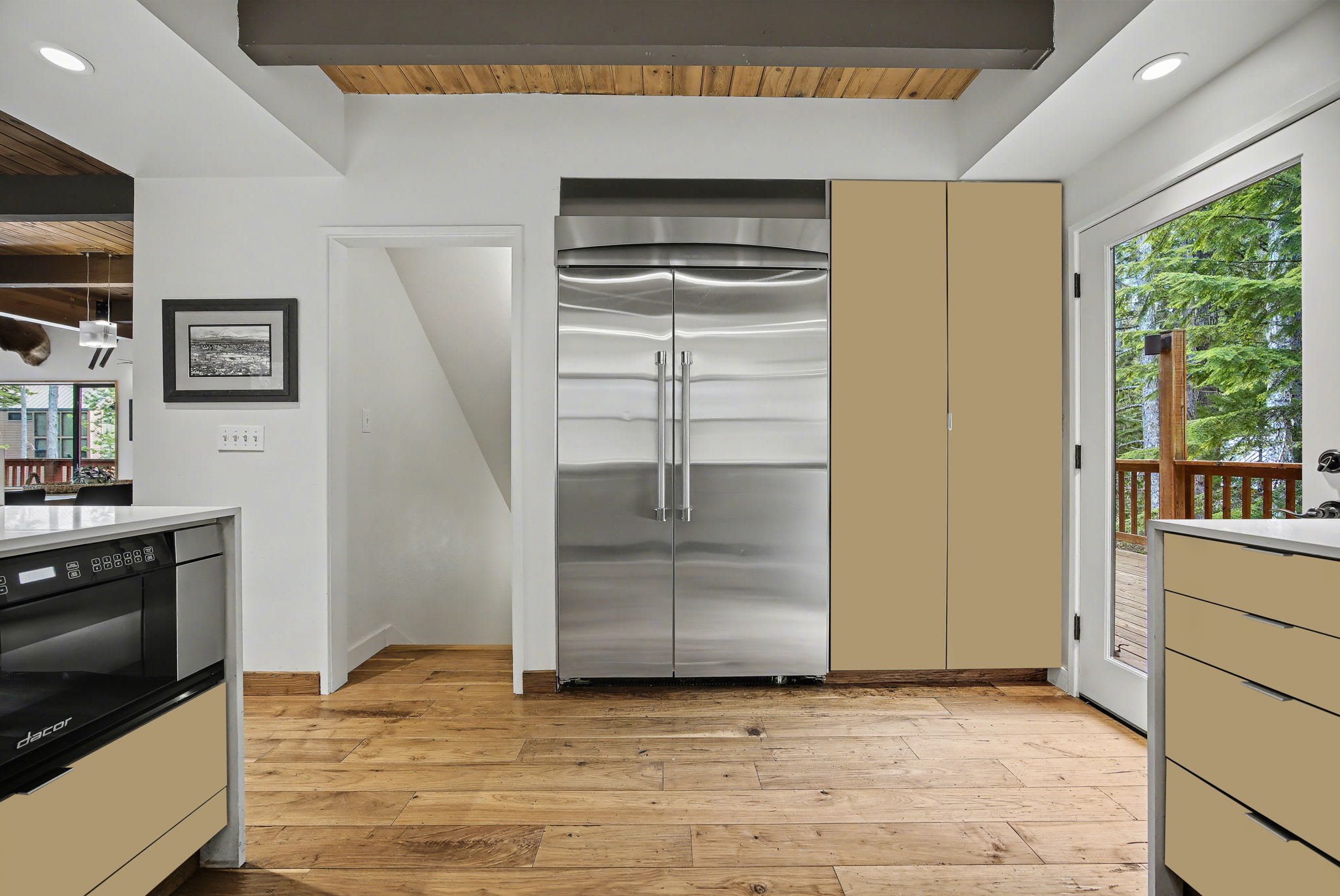 A wider view of the modern kitchen, showcasing a large stainless steel refrigerator and light cabinetry, with an open doorway leading to another area. Hardwood floors and a vaulted wooden ceiling maintain continuity with the rest of the house.