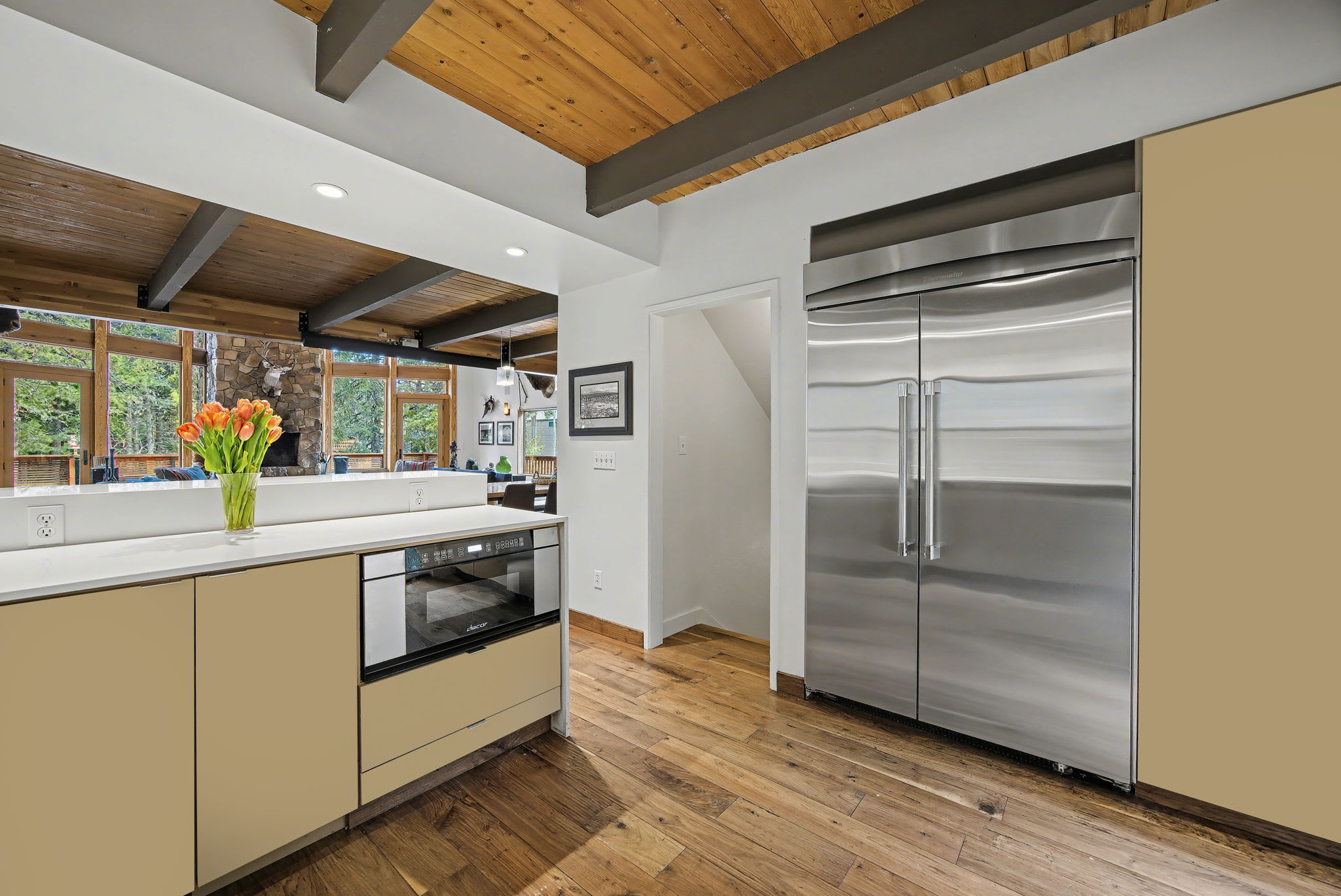 A modern kitchen area features light-colored cabinetry, white countertops, and stainless steel appliances including a large double-door refrigerator. Hardwood floors and a vaulted wooden ceiling extend from the adjacent living spaces.
