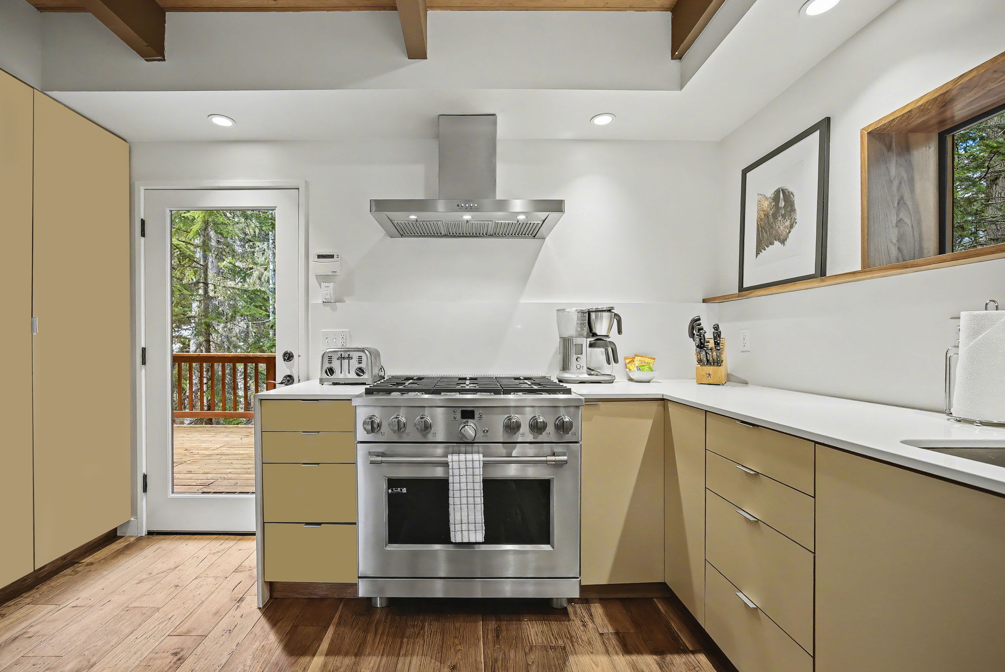 Modern kitchen with light wood cabinetry, white countertops, and stainless steel appliances including a gas range with a hood. A glass door leads to an outdoor deck surrounded by tall evergreen trees.