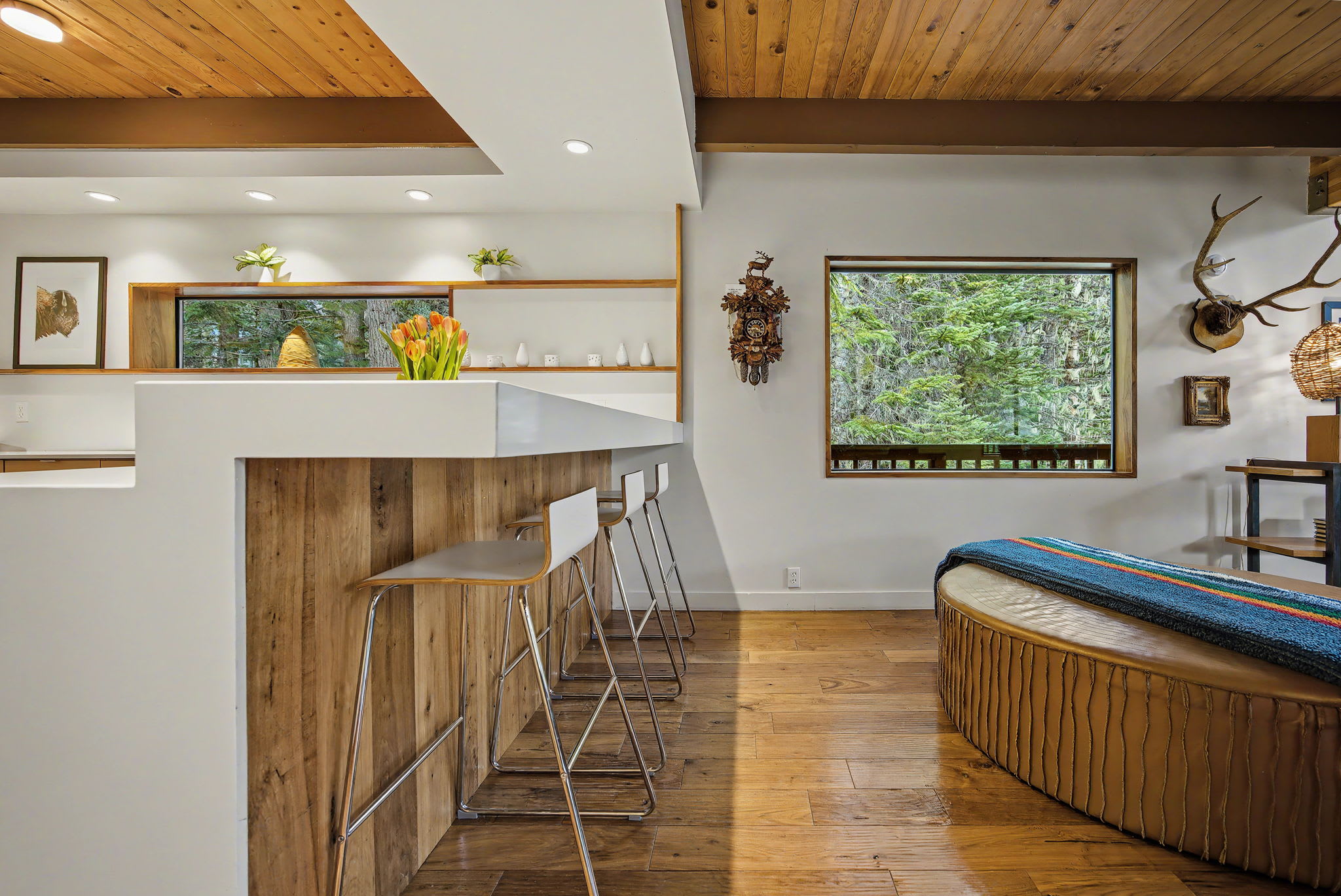 Open concept living area with a white bar counter and three modern bar stools, separating it from the main living space. A large window provides a picturesque view of the forest.