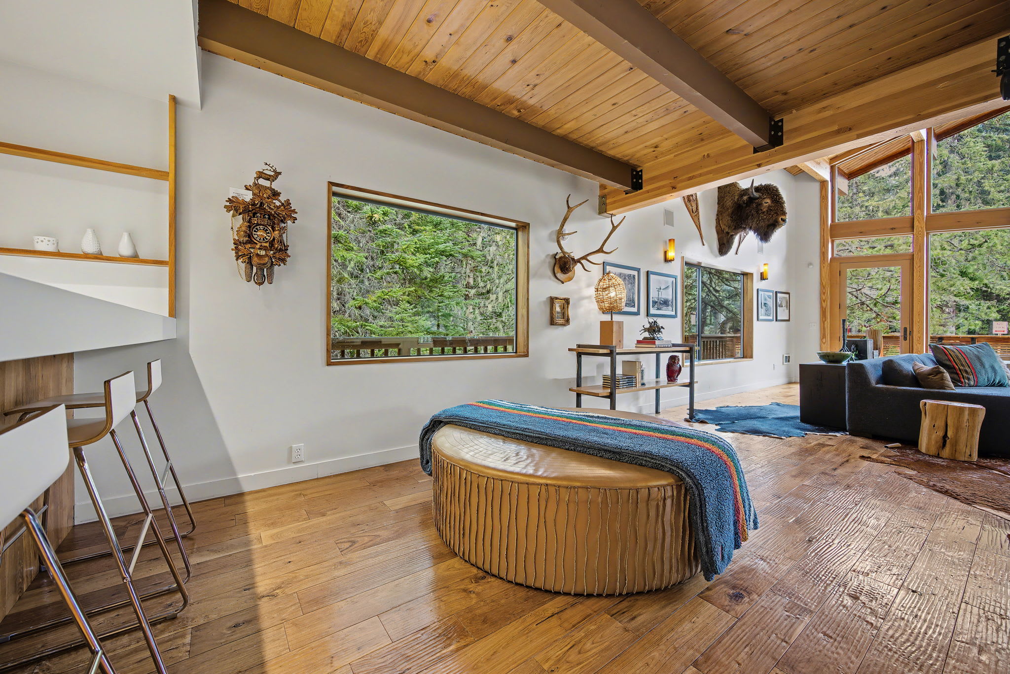 Spacious living room with warm hardwood floors, a large round leather ottoman, and a wood-paneled ceiling with exposed beams. Windows offer views of the surrounding evergreen trees.