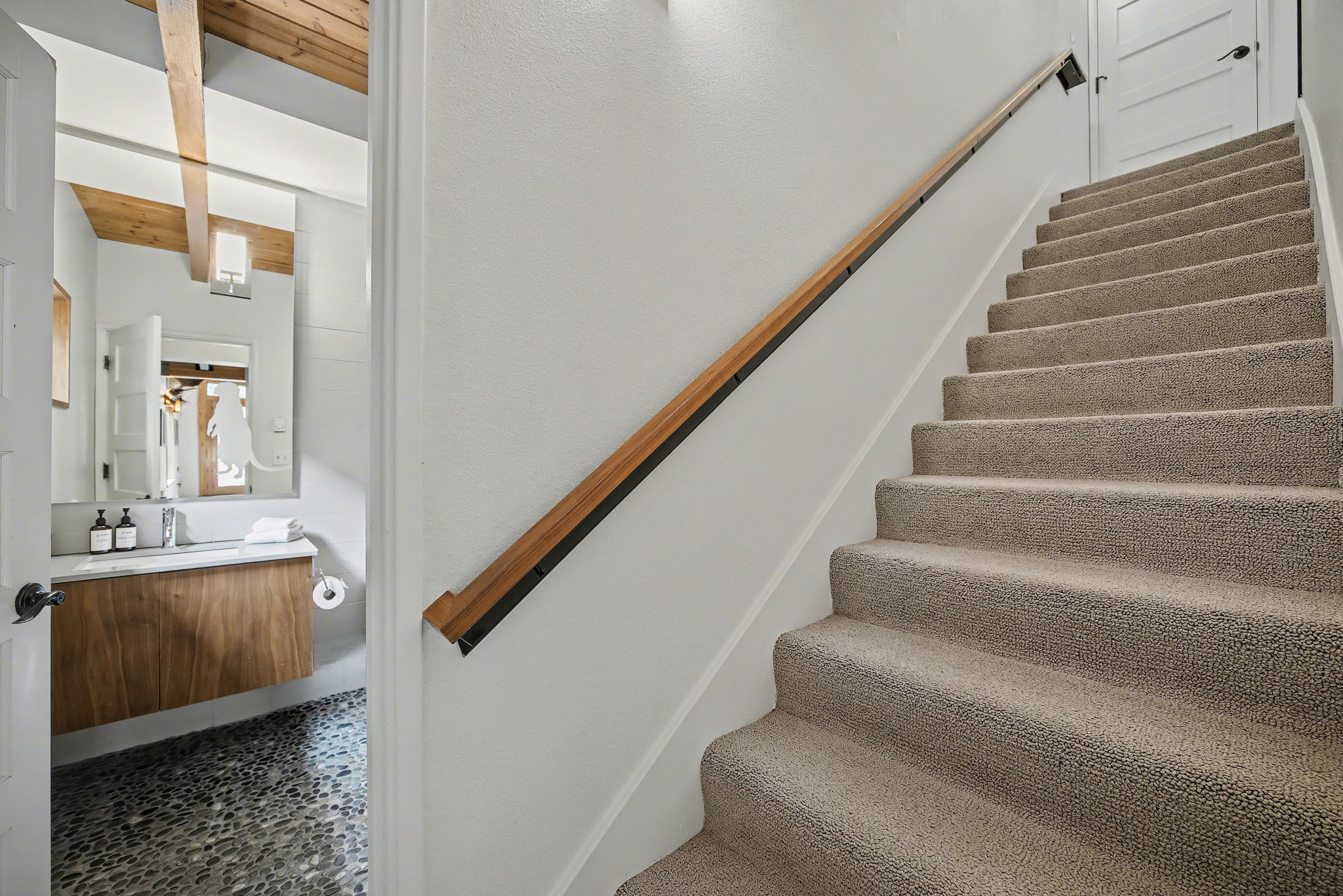 A carpeted staircase with a sleek wooden handrail leads upwards, offering a glimpse into a modern bathroom with a floating wooden vanity and a large mirror. The area is illuminated by a warm wooden beam ceiling.