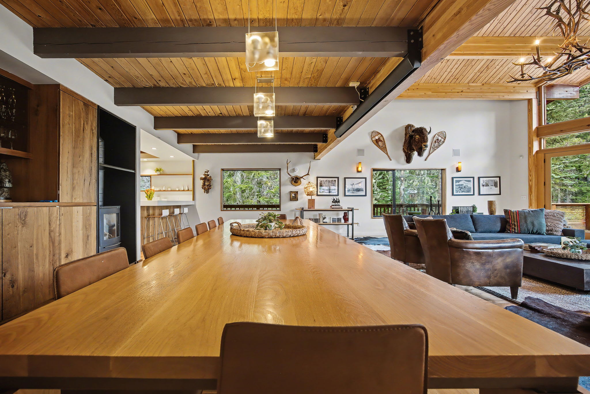 This view of the dining area from the end of the long wooden table highlights the open-concept layout leading to the living room. The space features a vaulted wooden ceiling, modern pendant lights, and a built-in bar with a wood-burning stove.