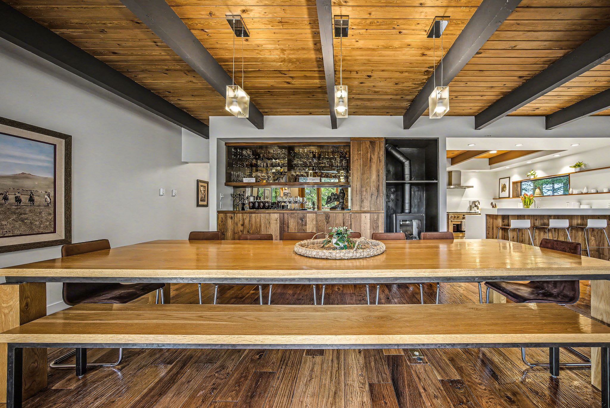 A spacious dining area features a long, light-wood dining table with benches and chairs, illuminated by modern pendant lights from a vaulted wooden ceiling. A built-in bar with mirrored shelves and a wood-burning stove are visible in the background.