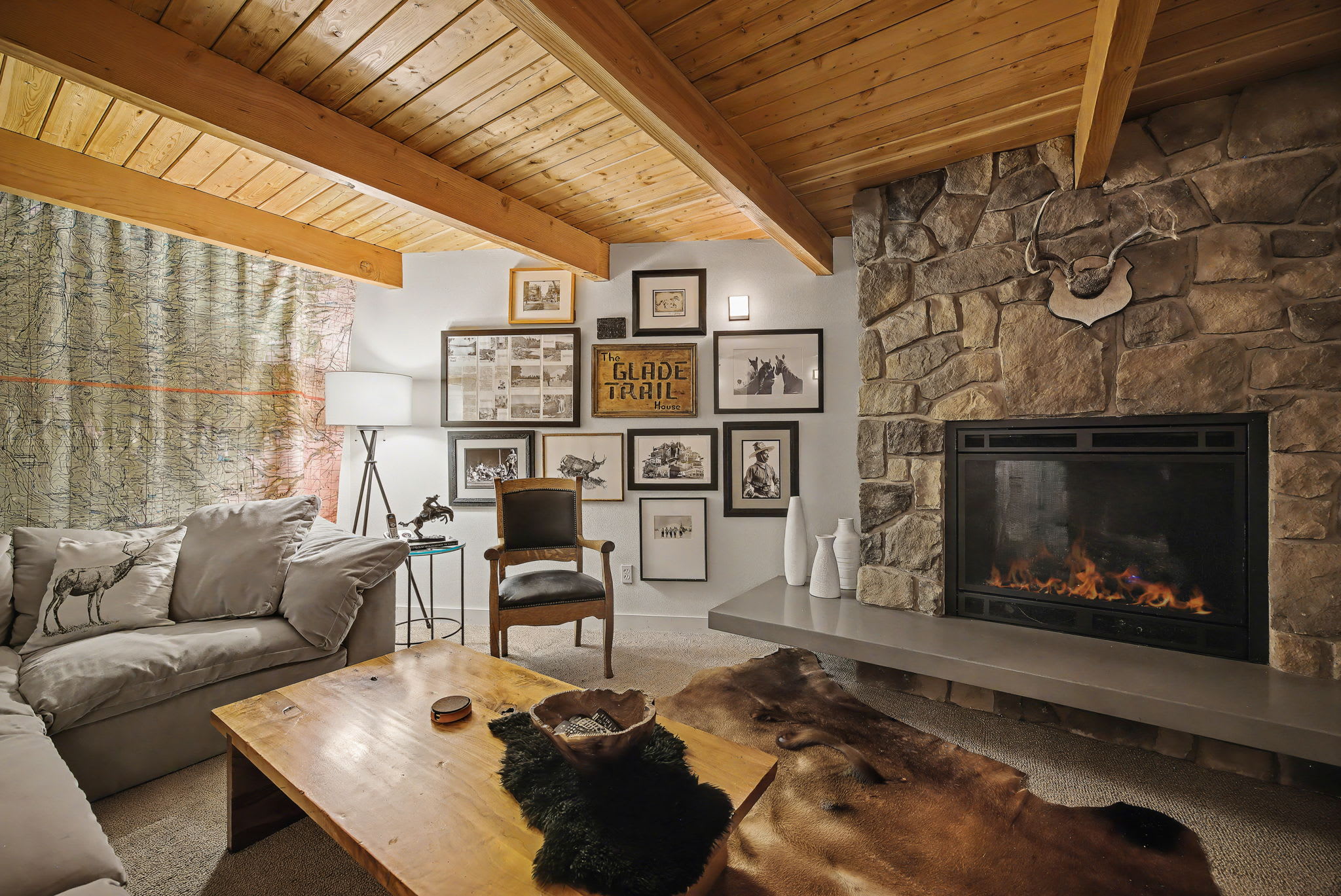 A cozy living room featuring a large L-shaped sofa, a rustic wooden coffee table, and a prominent stone fireplace with a modern hearth. The room is adorned with framed artwork on the walls and a warm wooden beam ceiling.