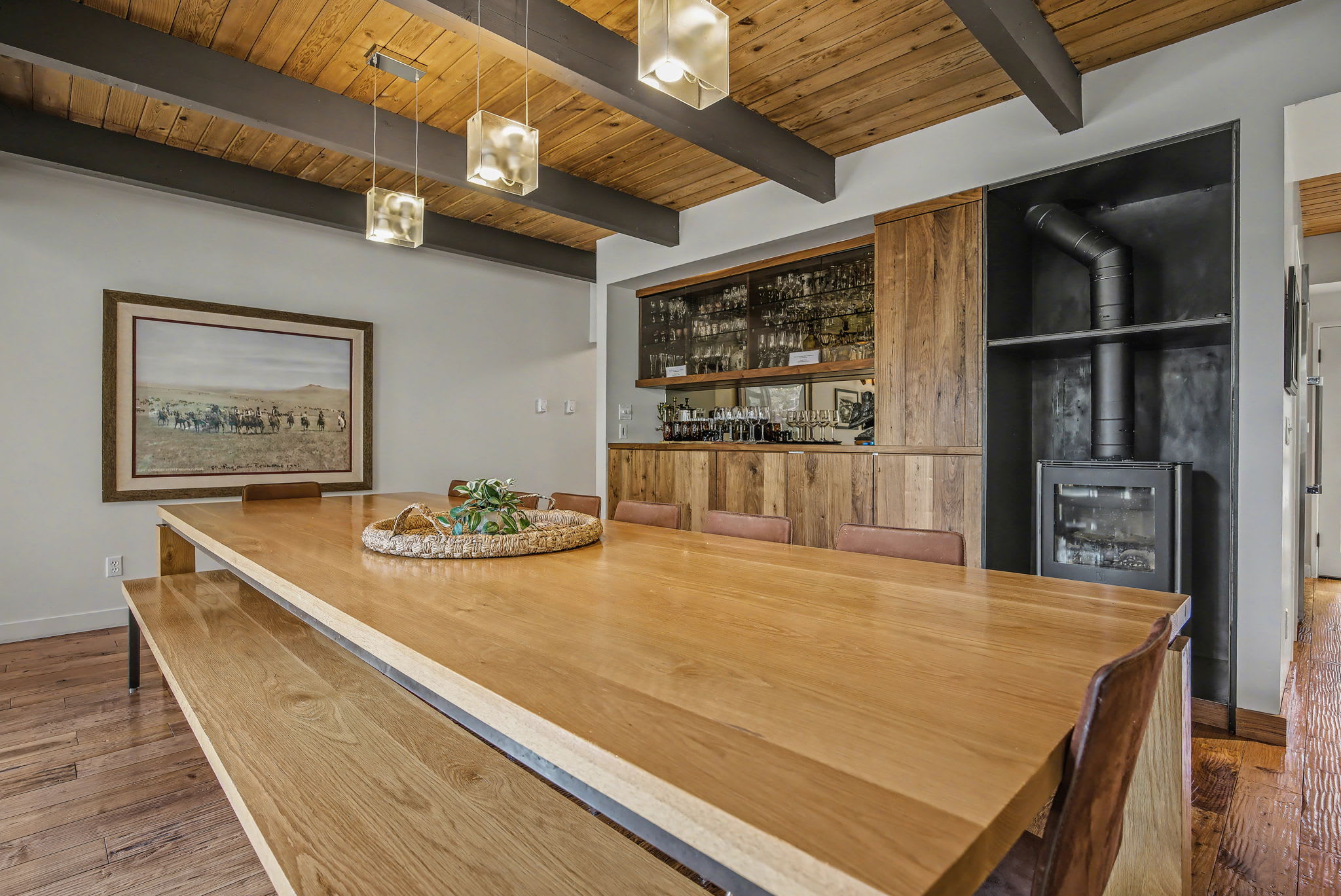 A close-up of the long wooden dining table with benches and chairs, showcasing its rustic-modern design. The background reveals a built-in bar with mirrored shelves and a wood-burning stove, all under a vaulted wooden ceiling.