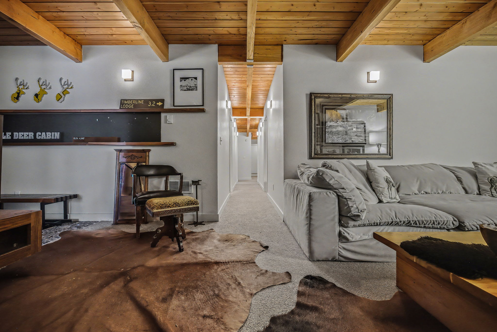 Cozy living area featuring a vaulted wooden beam ceiling, light gray walls, and two cowhide rugs on carpet, with a large light-colored sofa and a dark leather chair facing a hallway.