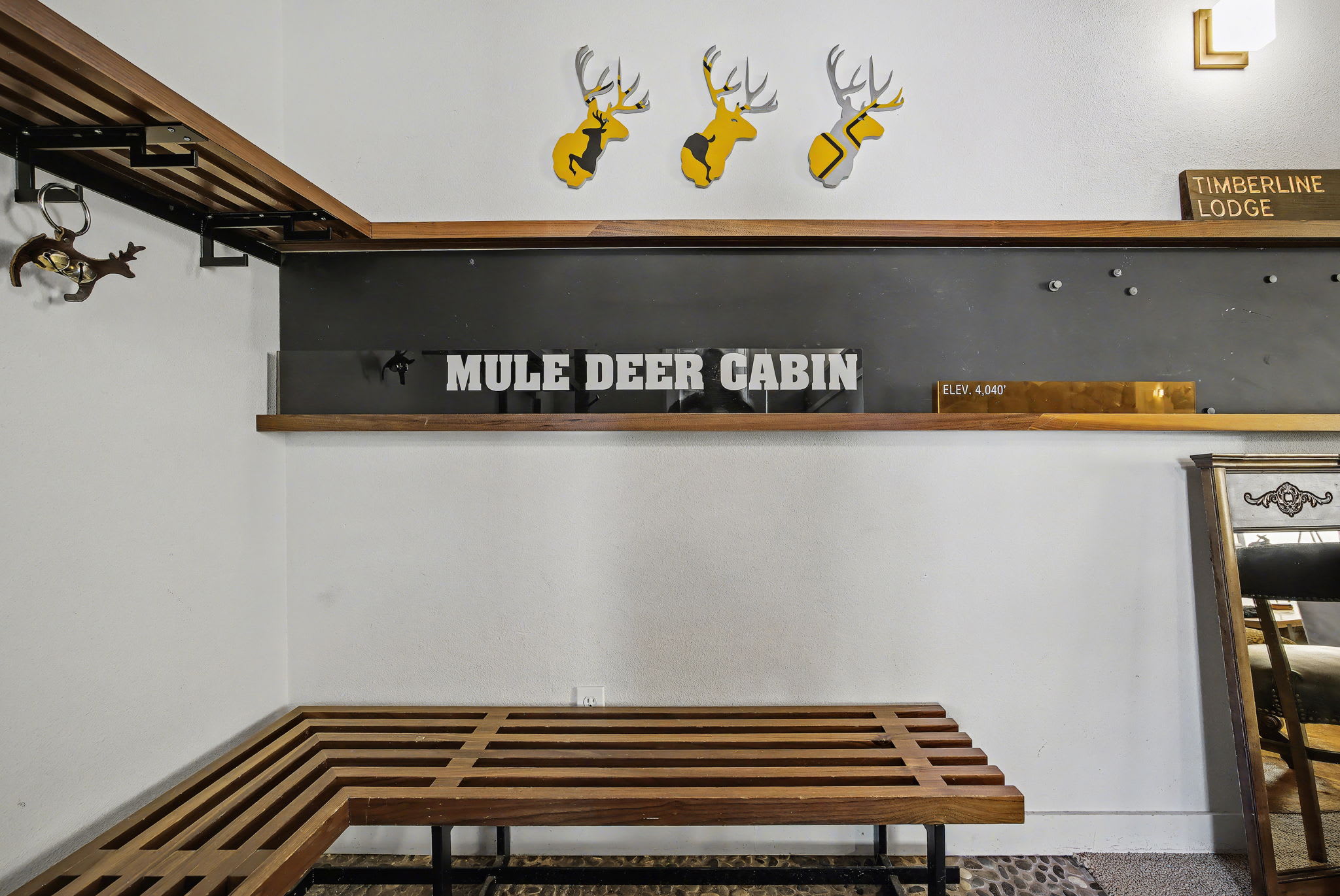 Entryway wall with a 'MULE DEER CABIN' sign and 'TIMBERLINE LODGE' directional sign, featuring three yellow deer head decorations and a built-in wooden slatted bench.