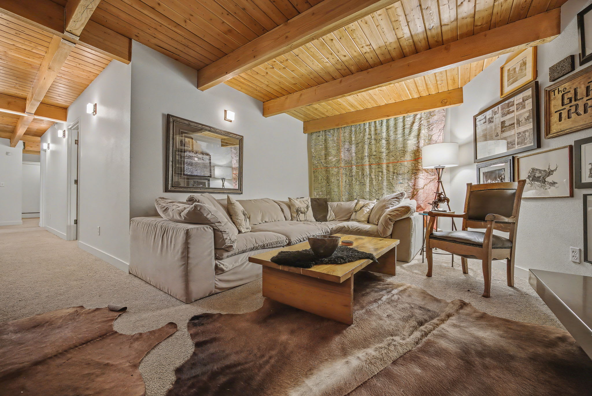 A spacious living room with a comfortable L-shaped sofa, a wooden coffee table, and a stone fireplace, creating a warm and inviting atmosphere. The room features a wooden beam ceiling, a gallery wall of framed art, and cowhide rugs on the carpeted floor.
