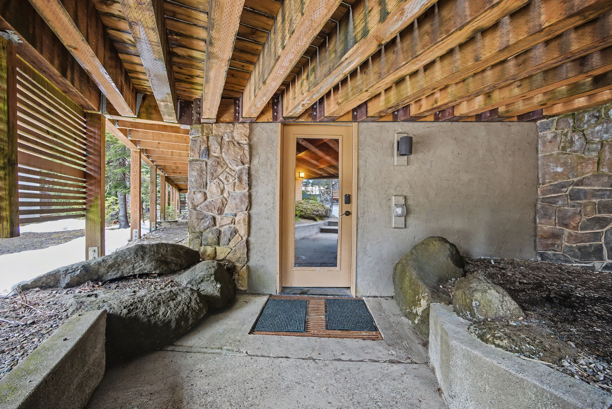 Front entrance door with a glass pane, set into a stone and stucco facade, located under a large wooden deck structure, with a concrete path flanked by large rocks.