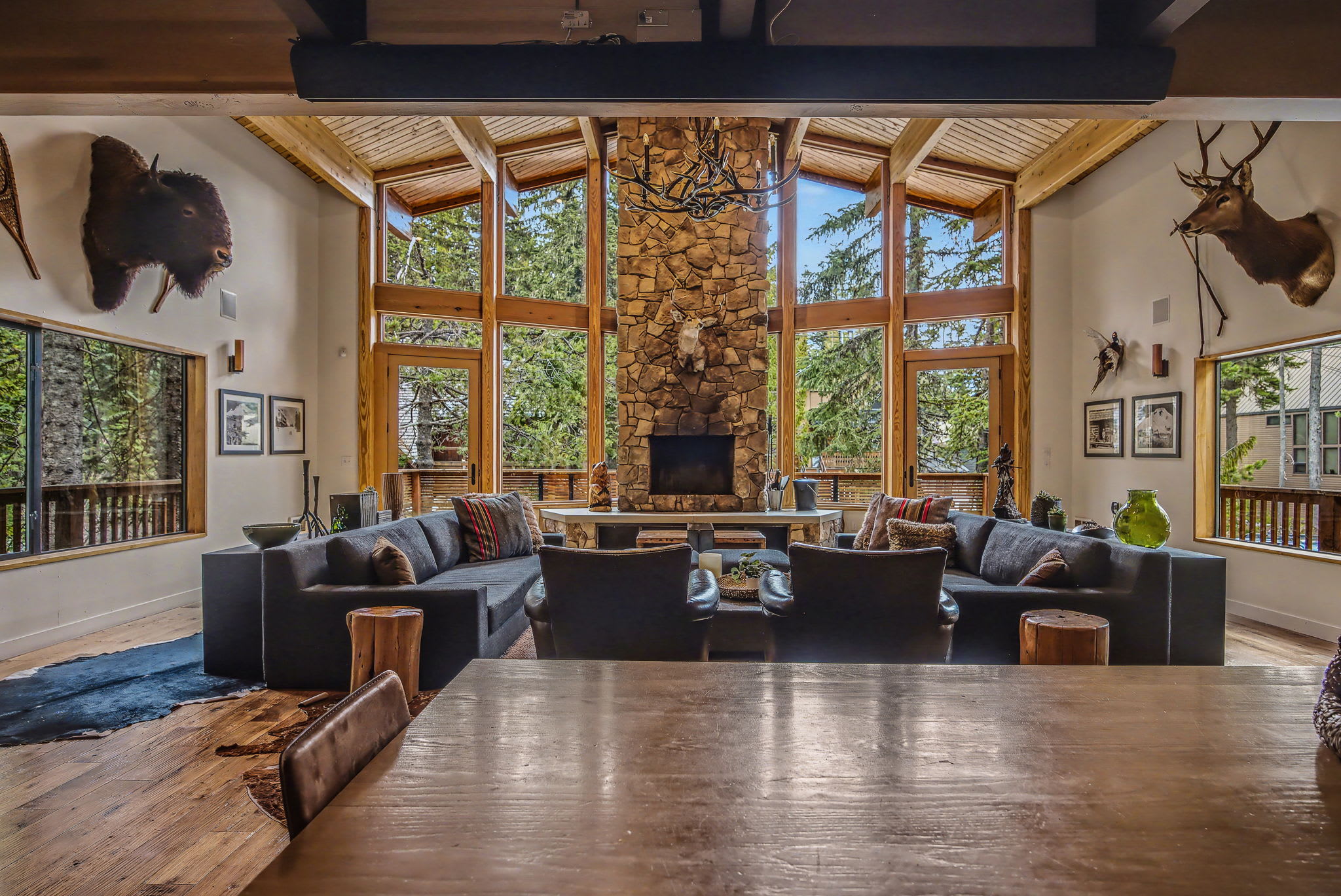 A grand living room features a central stone fireplace and floor-to-ceiling windows offering panoramic views of the surrounding forest. Multiple dark sofas and armchairs are arranged around a large coffee table under a vaulted wooden ceiling.