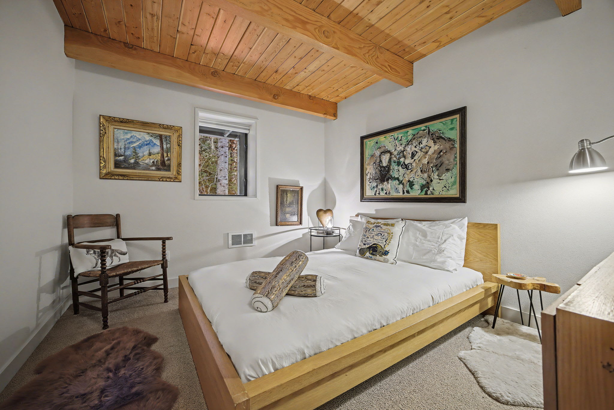 A cozy bedroom with a wooden bed frame, white bedding, and a large abstract painting above the headboard. The room features a warm wooden beamed ceiling, carpeted floor, and a small window overlooking trees.