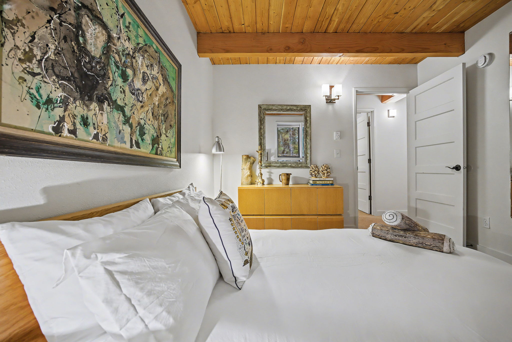 A different perspective of the bedroom, showcasing a wooden dresser with a decorative mirror and sconce lighting, and a large abstract painting above the bed. A white door leads to another area, and the wooden beamed ceiling continues throughout.