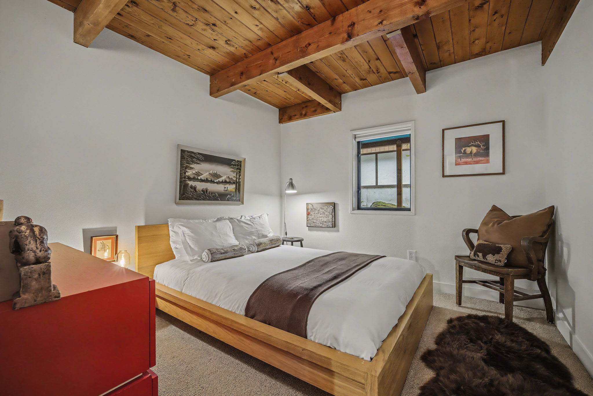 A bright bedroom featuring a wooden bed frame with white bedding and a brown throw, complemented by a vibrant red dresser. The room has a carpeted floor, a wooden beamed ceiling, and two framed artworks on the white walls.