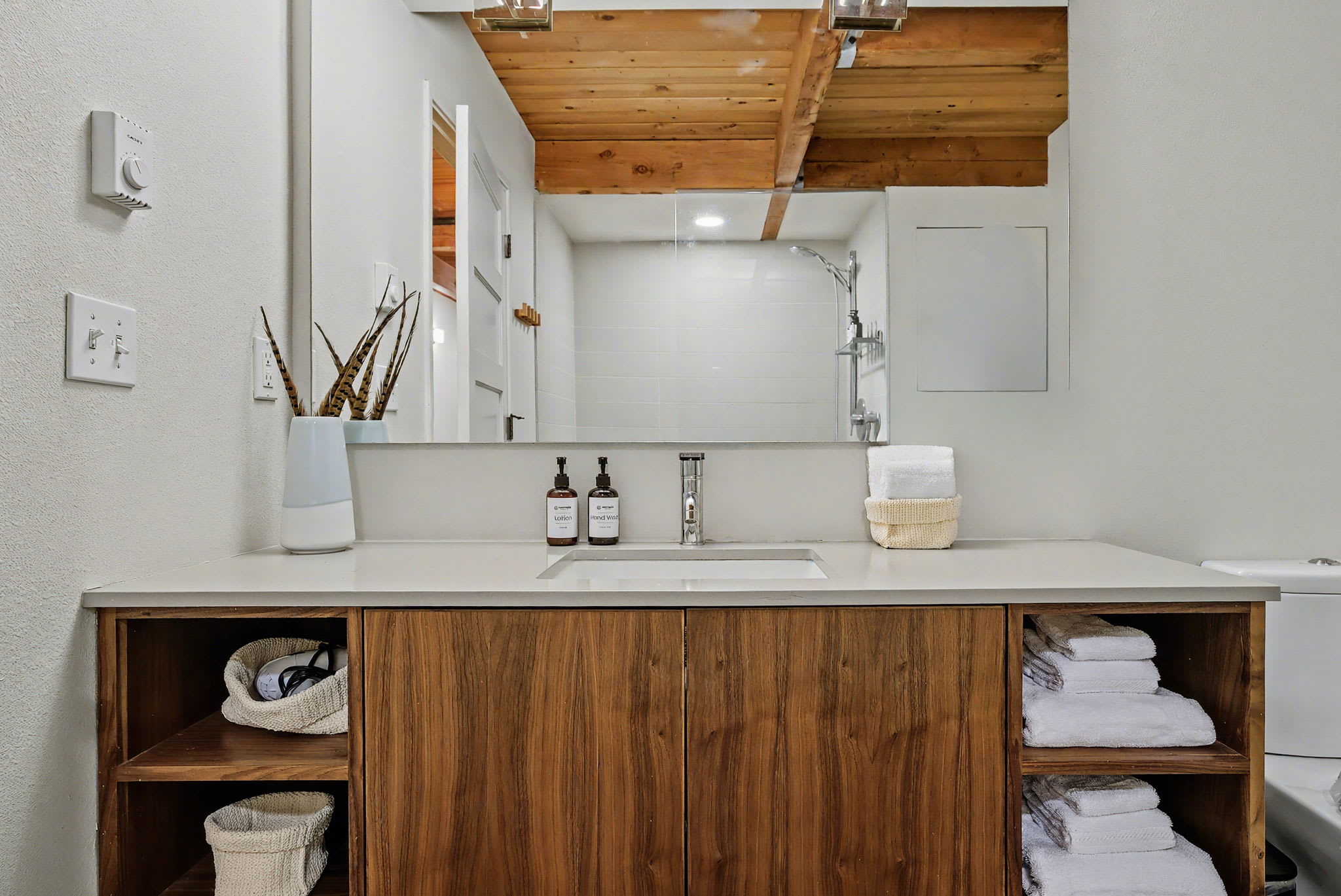 Modern bathroom vanity with a single sink set in a light countertop, a wooden cabinet with open shelving, a large mirror, and neatly folded white towels, under a wooden ceiling.