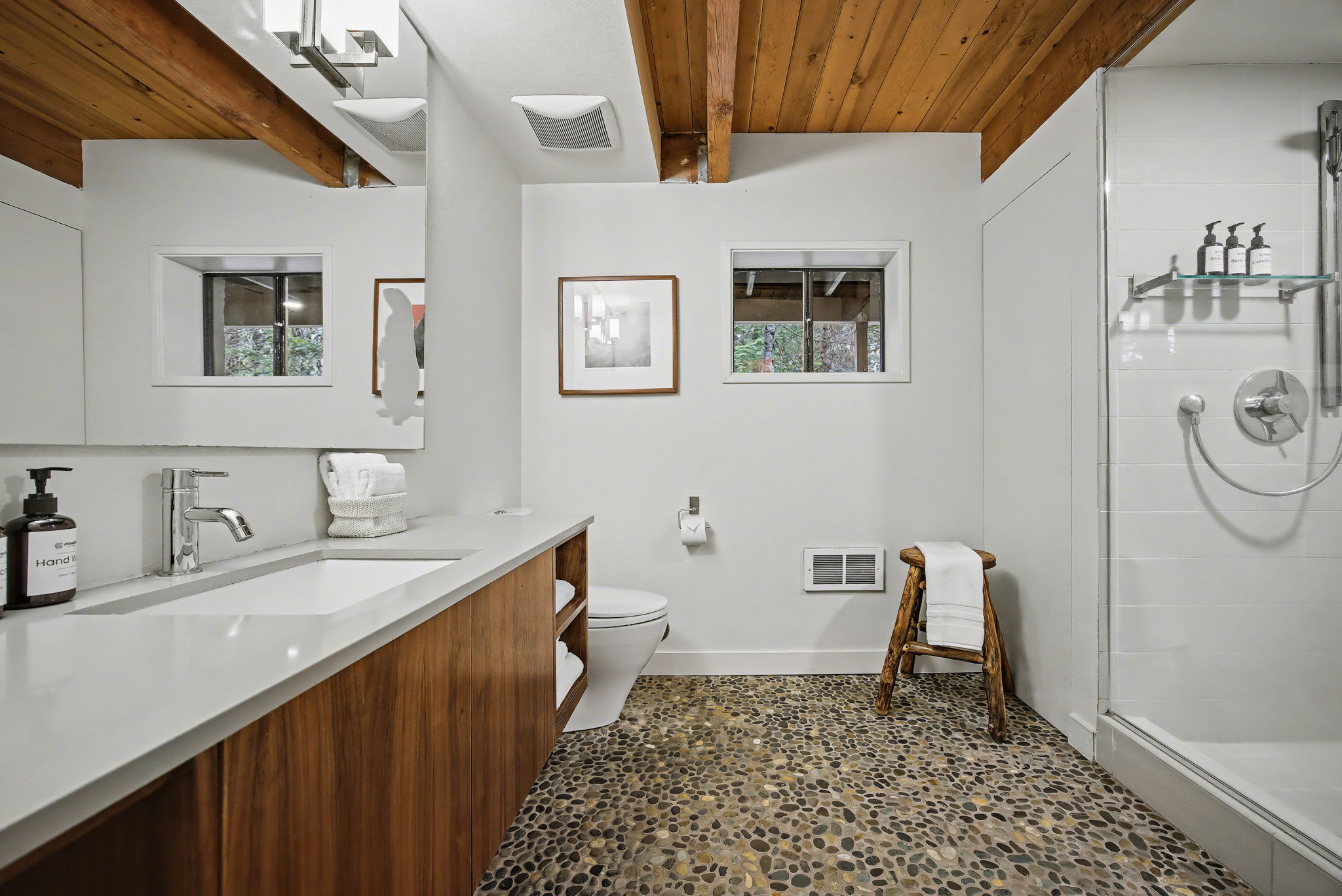 A modern bathroom featuring a unique pebble-tiled floor, a light wood vanity with a white countertop, and a walk-in shower with a glass enclosure. The room has white walls and a warm wooden beamed ceiling.
