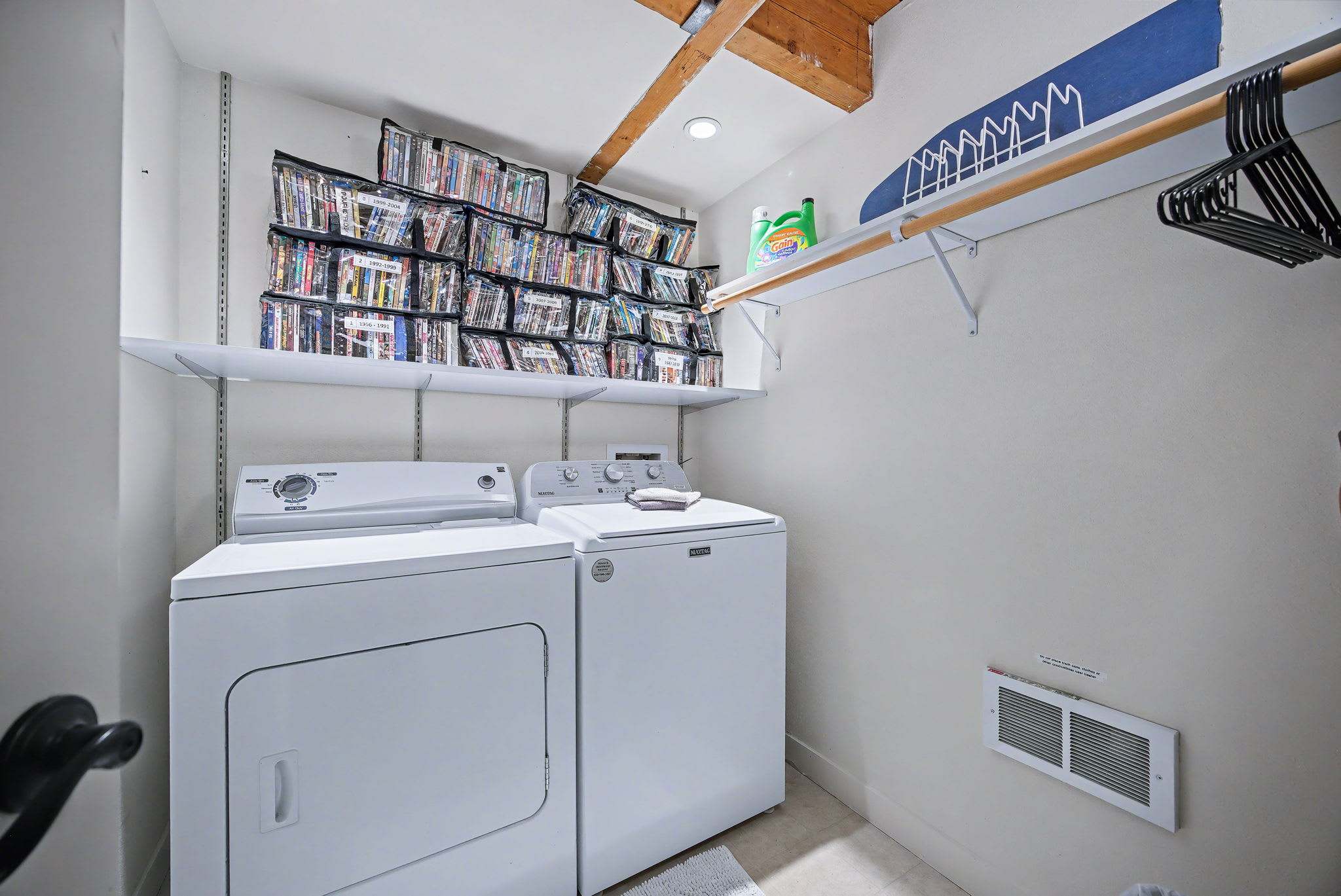 A functional laundry room equipped with a white washer and dryer, overhead shelving filled with DVDs, and a wall-mounted ironing board. The room has white walls and a wooden beamed ceiling.