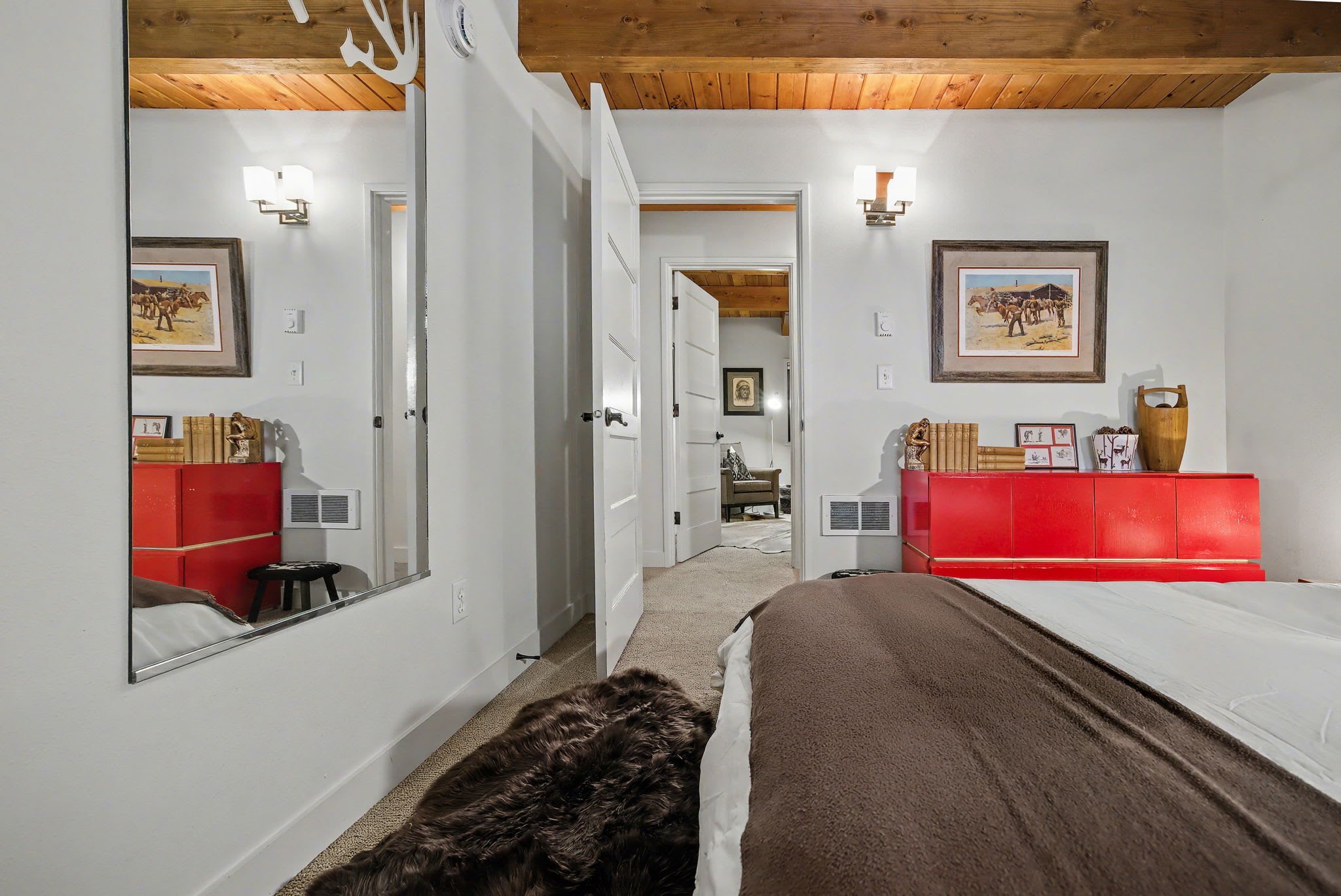 A view of the bedroom highlighting a full-length mirror reflecting the red dresser and a framed artwork. The room features a wooden beamed ceiling and a white door leading to an adjacent hallway.