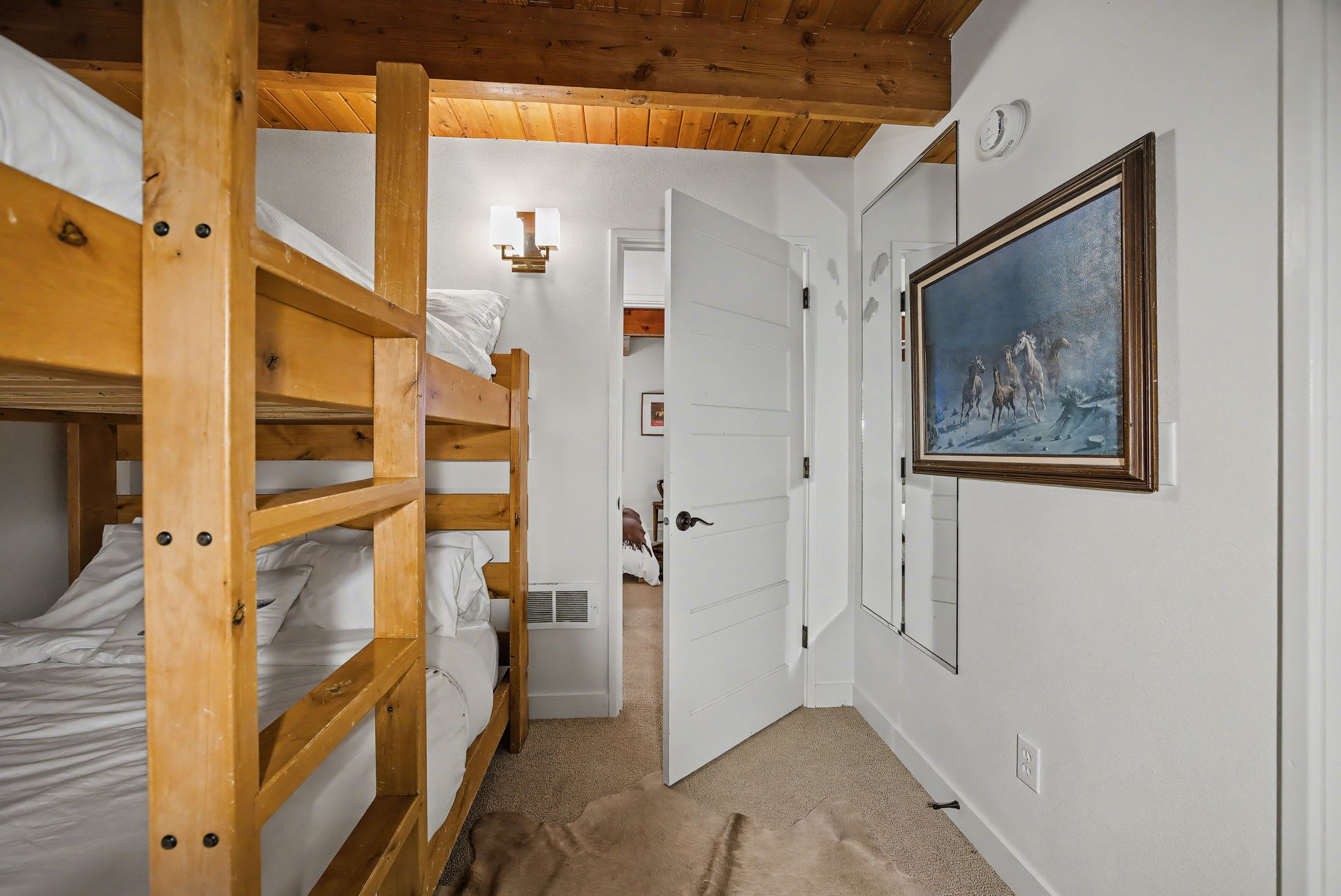 Bedroom with sturdy wooden bunk beds and white bedding, featuring a doorway leading to another room, a mirror, and a framed painting of horses on the wall, under a wooden ceiling.