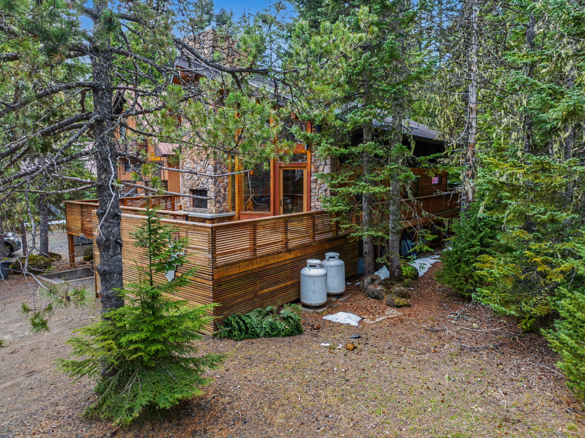 An aerial view of the house's side, revealing its stone facade, a wooden deck, and two large propane tanks nestled among dense pine trees. The property is surrounded by a natural forest setting.