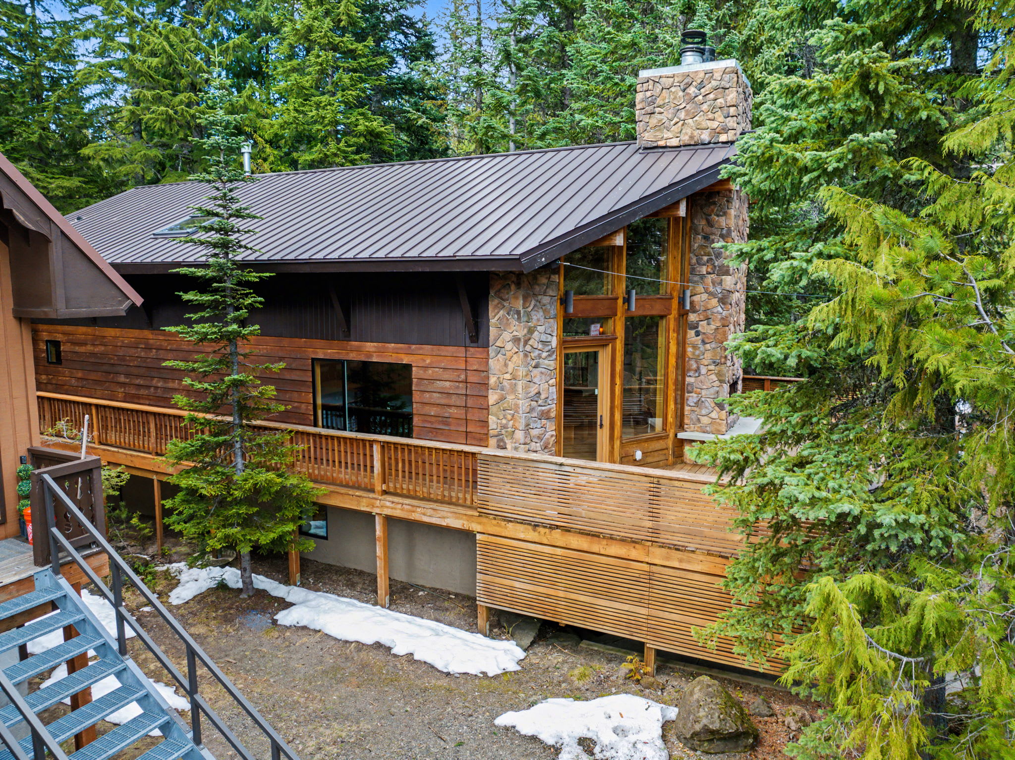 The back of a rustic chalet features a large wooden deck with slatted railings, a prominent stone chimney, and expansive windows, surrounded by tall evergreen trees and patches of snow on the ground.