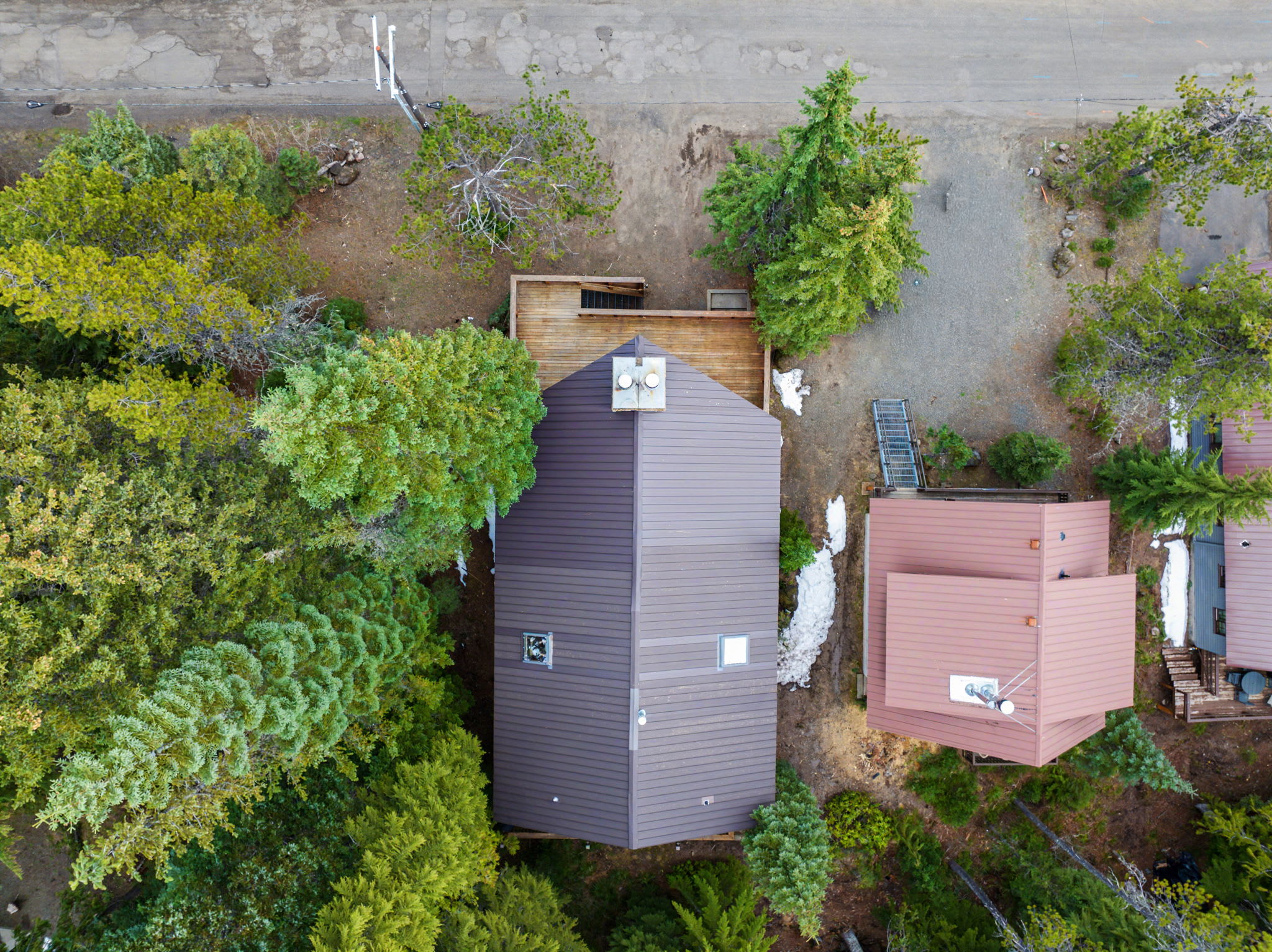An aerial view showcases the property's dark metal roof and chimney, nestled among dense evergreen trees, with a gravel driveway and a paved road visible nearby, indicating a secluded yet accessible location.