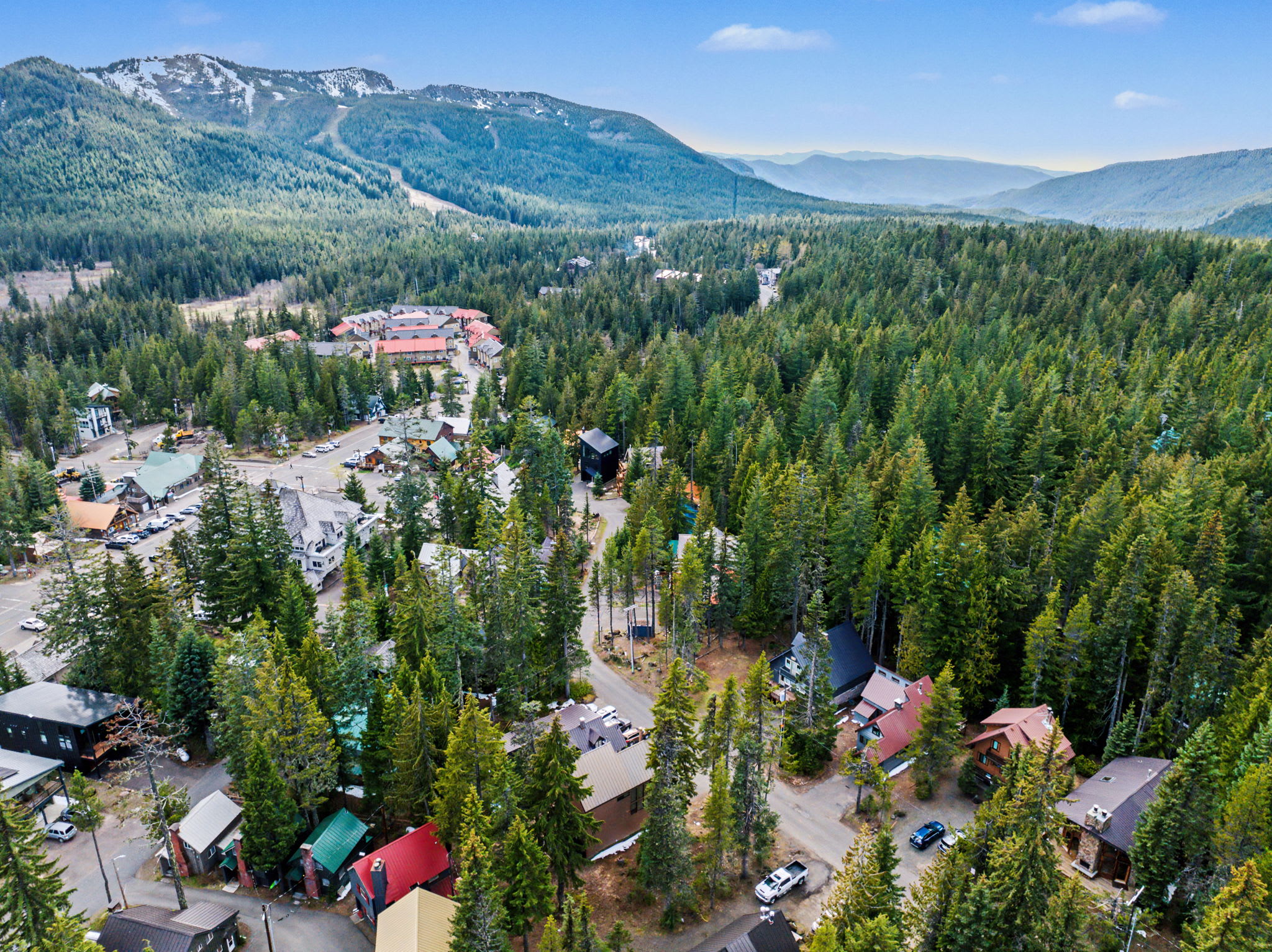 A panoramic aerial view captures a charming mountain community with numerous cabins and chalets nestled within a vast evergreen forest, with majestic snow-capped mountains in the distance under a clear blue sky.