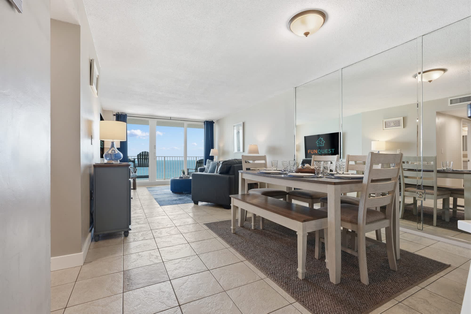 Dunes E1702 – Living room and dining area with ocean view, featuring a table, blue accents, and sliding glass doors.
