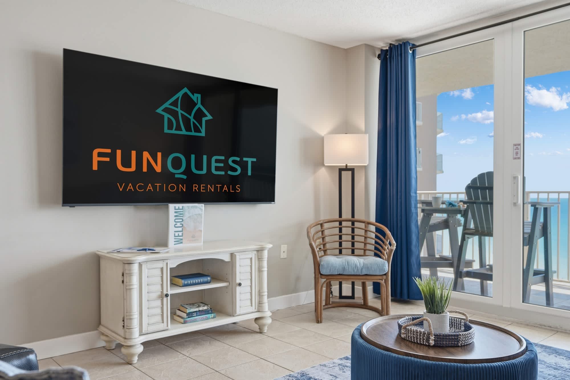 Dunes E1702 – Living room with a large TV, blue curtains, and balcony view of the ocean.