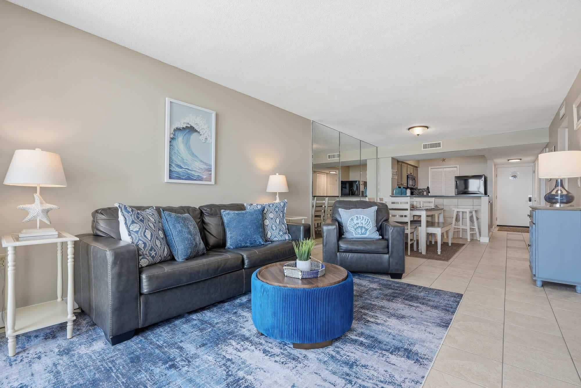 Dunes E1702 – Living room with gray leather sofa, blue accents, round coffee table, and open kitchen area visible.
