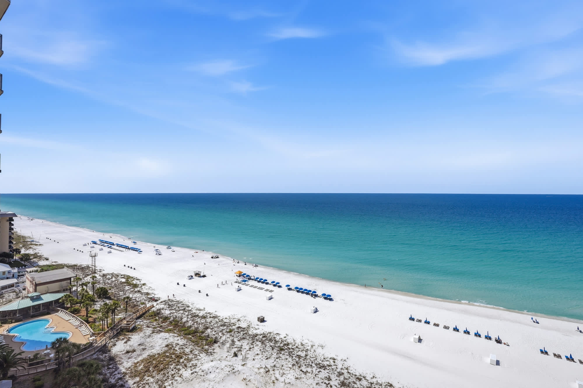 Dunes E1702 – View of a beach with white sand, blue umbrellas, and a pool area below.