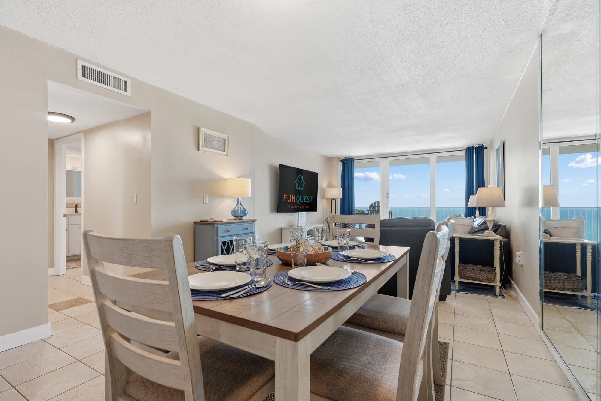 Dunes E1702 – Dining area with table set, view of ocean from large windows, living space with sofa and TV.