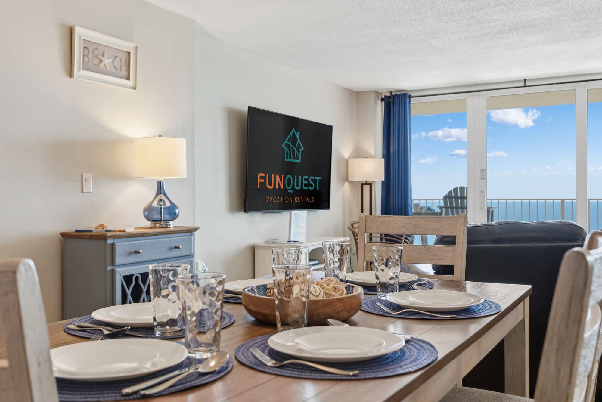 Dunes E1702 – Dining area with set table, large TV, and ocean view through sliding glass doors.