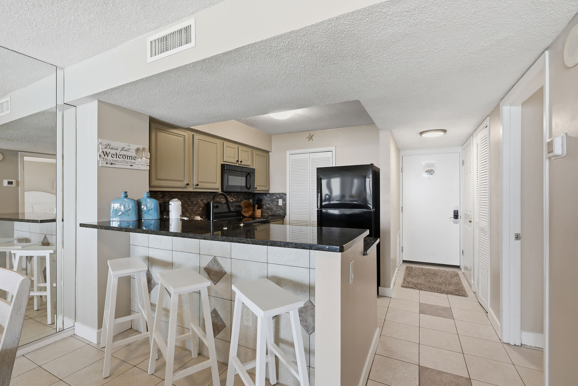 Dunes E1702 – Kitchen area with black appliances, granite counter, and white bar stools; entrance visible in background.