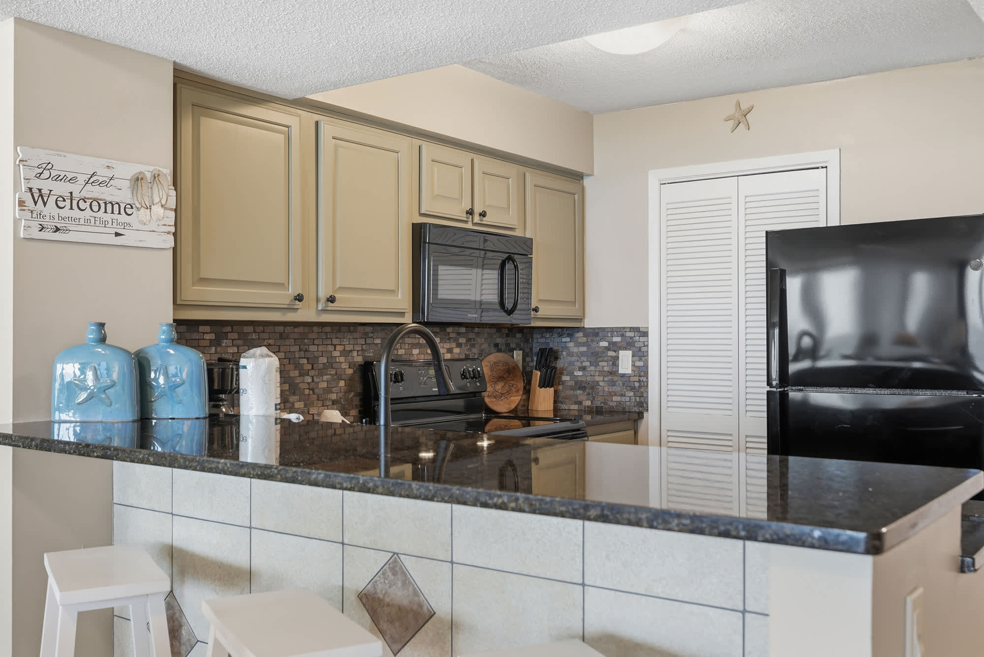 Dunes E1702 – Kitchen with granite countertop, black appliances, and beige cabinetry, featuring decorative blue bottles and wooden utensils.