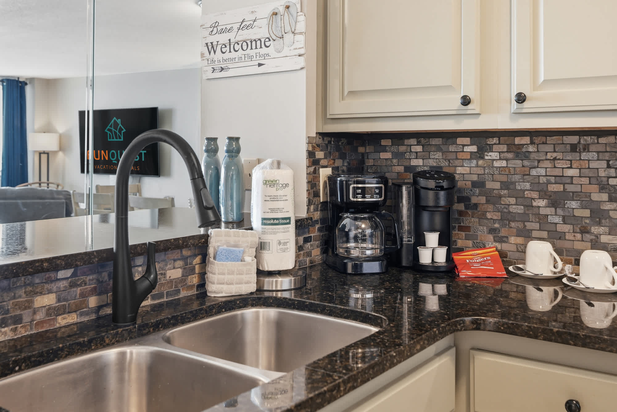 Dunes E1702 – Modern kitchen with dark countertops, coffee maker, utensils, and decorative vases; small living area visible in the background.