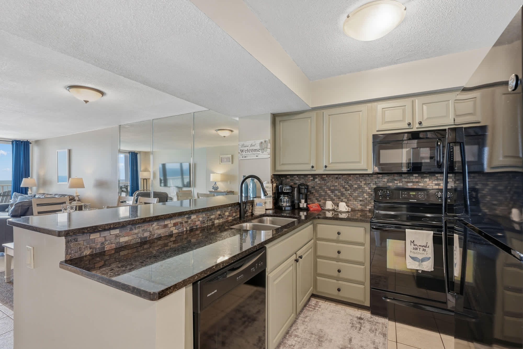 Dunes E1702 – Modern kitchen with granite countertops, black appliances, and a view of the living area with blue curtains.