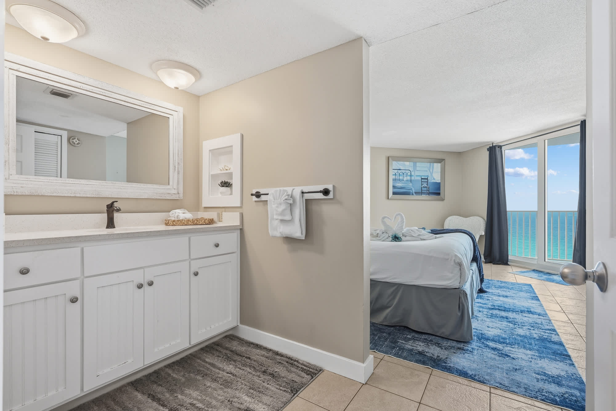 Dunes E1702 – Bathroom with sink and mirror; view of ocean from bedroom with bed and blue rug.