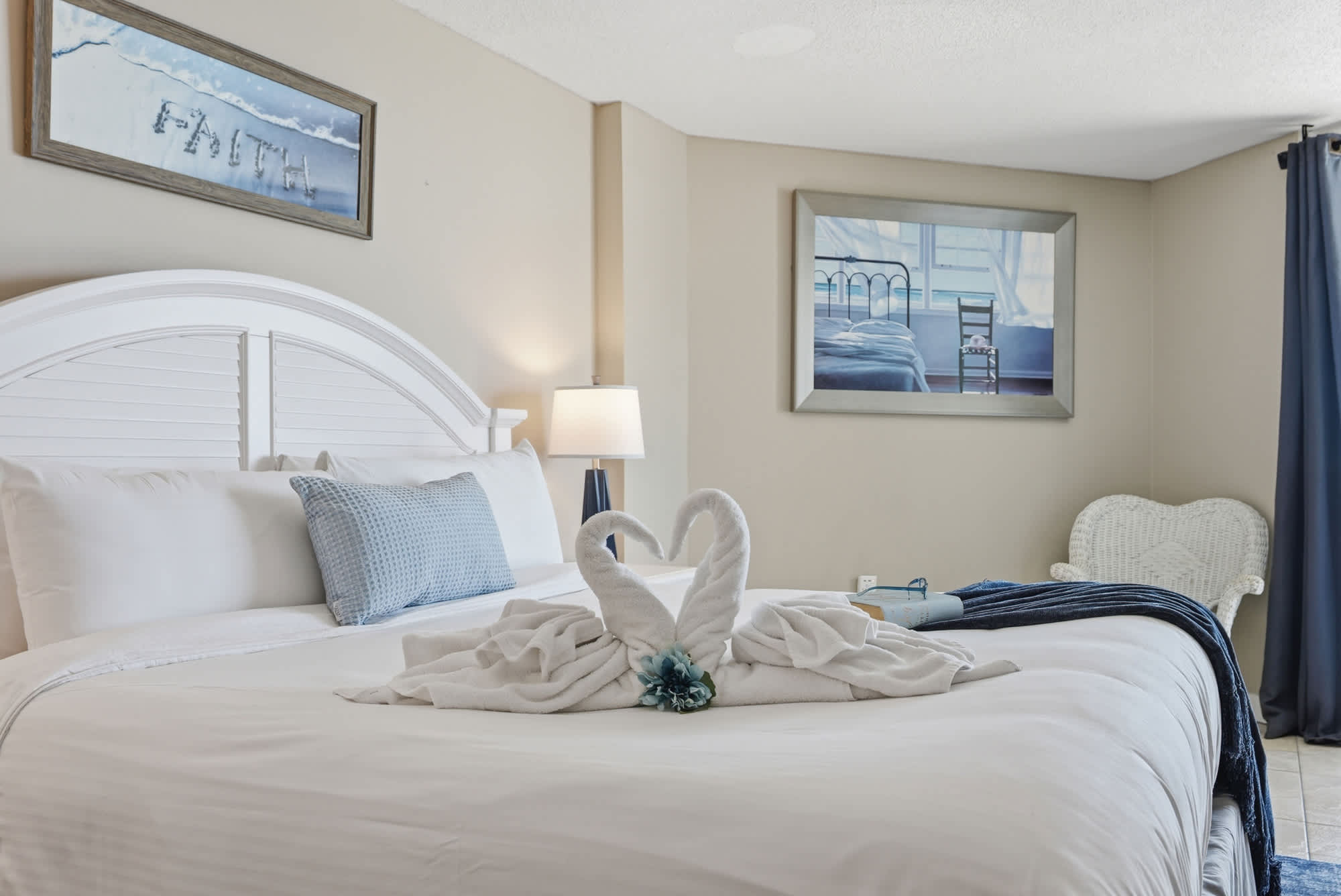 Dunes E1702 – Cozy bedroom with a king bed, lamp, artwork, and towel swans on the bed.