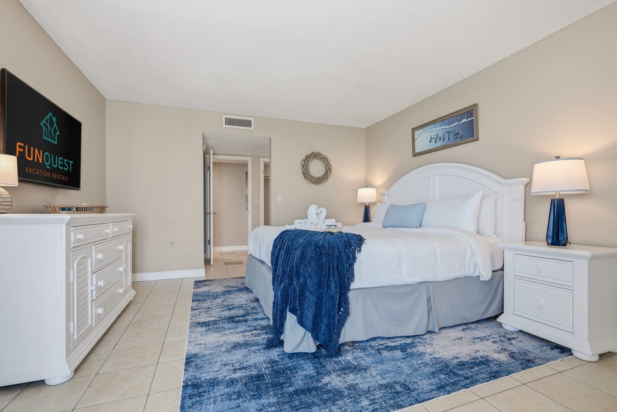 Dunes E1702 – Cozy bedroom with a king-size bed, white furniture, illuminated lamps, and a blue rug.