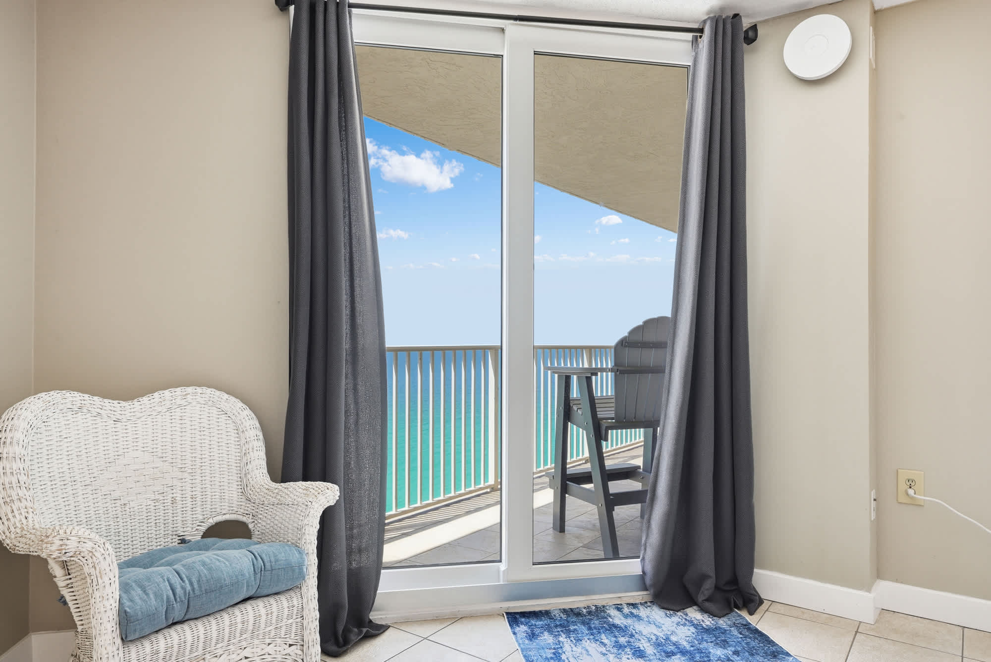 Dunes E1702 – Cozy corner with a wicker chair, sliding glass door, and balcony view of ocean and sky.