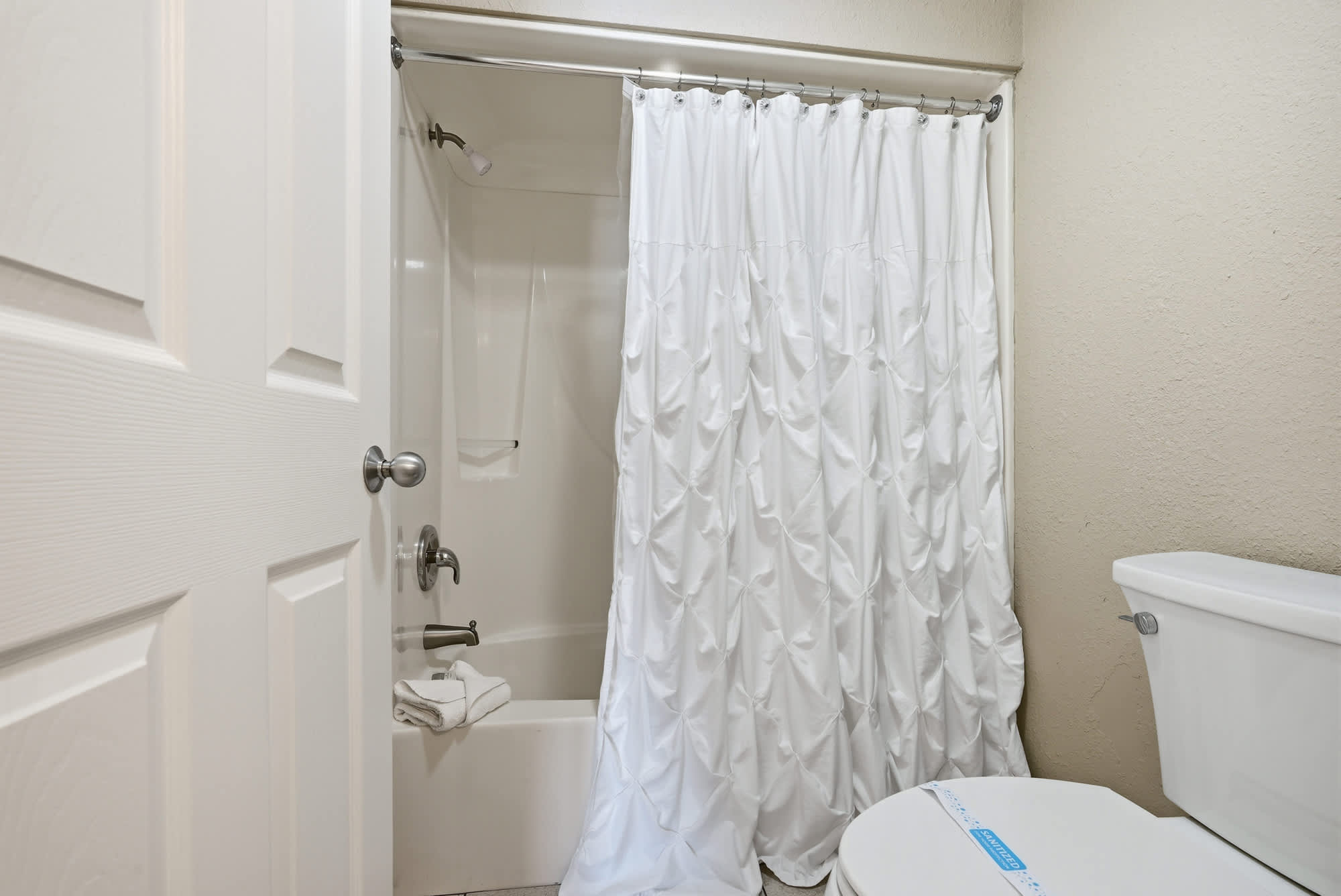 Dunes E1702 – Bathroom with a shower, white shower curtain, toilet, and towels on the tub edge.